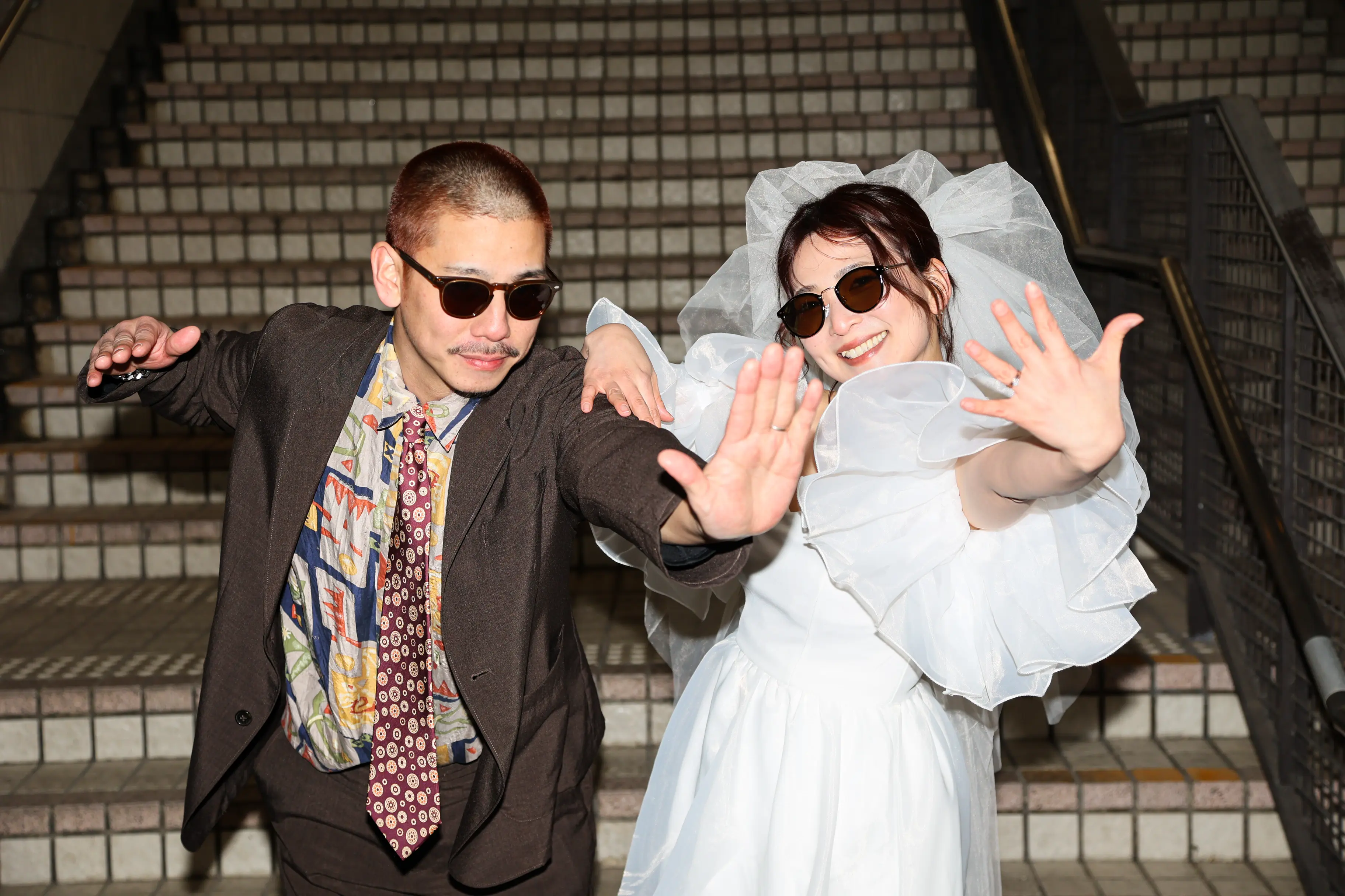 金沢の階段の前で両手を広げおどけるサングラス姿の白いドレスの新婦とブラウンのスーツの新郎
