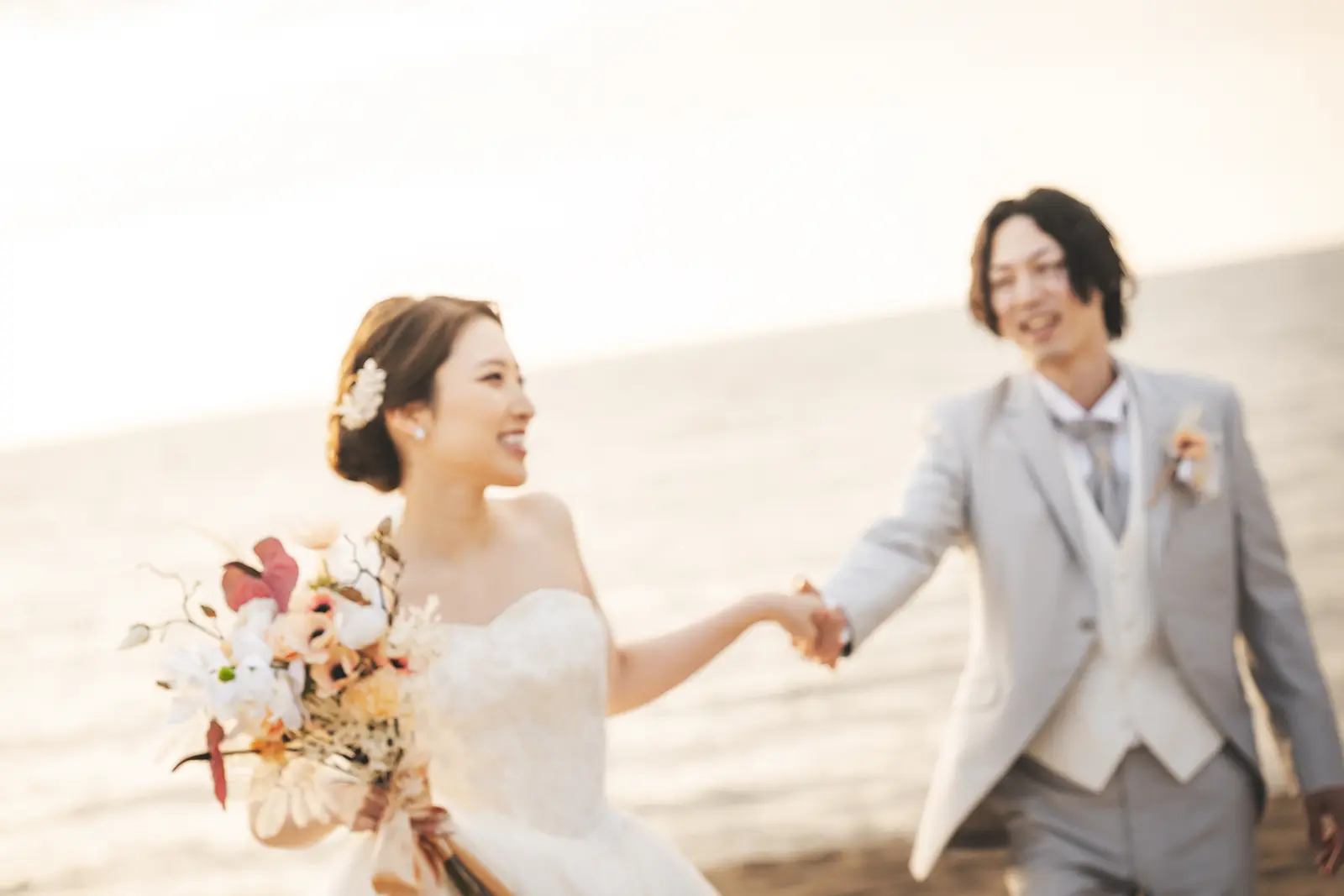 海辺で手をつないで歩く洋装の新郎新婦