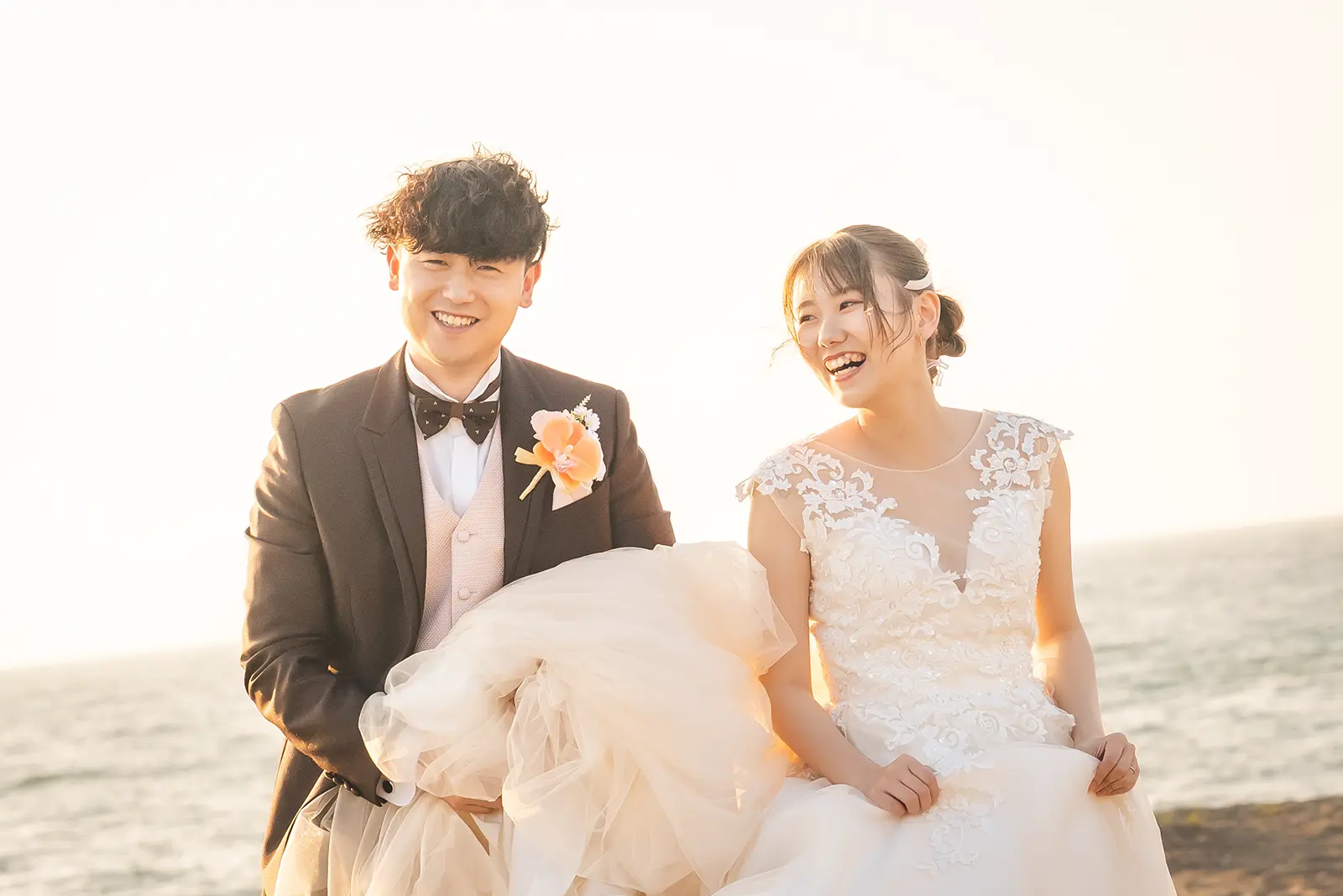 夕暮れの海辺で笑顔を見せる洋装の新郎新婦