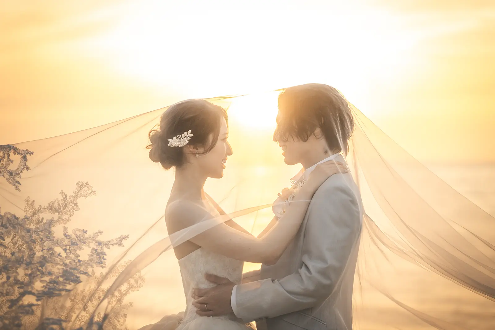 夕陽の海辺でベールに包まれる洋装の新郎新婦