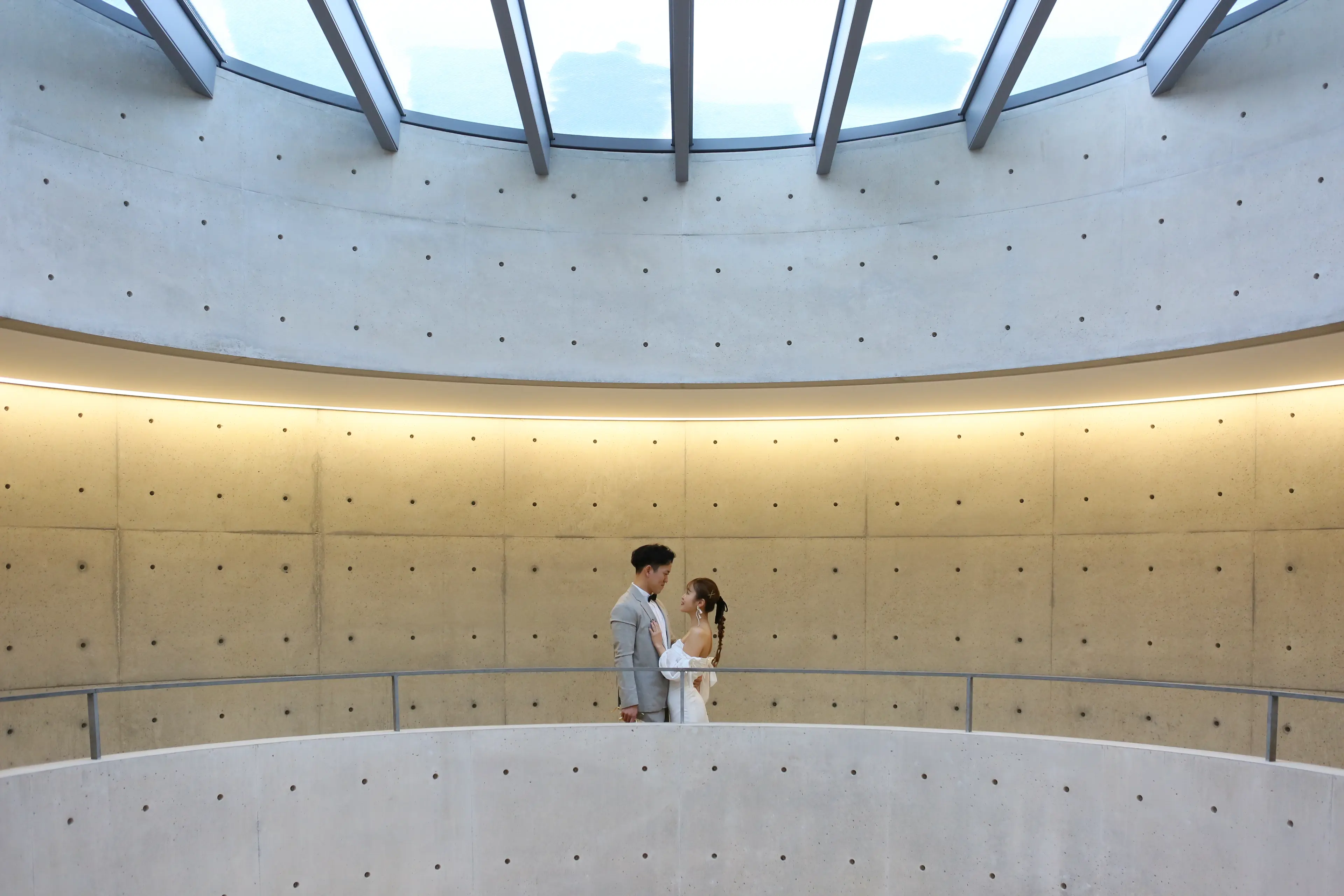西田幾多郎記念哲学館の円形ホワイエで、トップライトから光が差すコンクリートの曲面壁を背に向かい合う新郎新婦