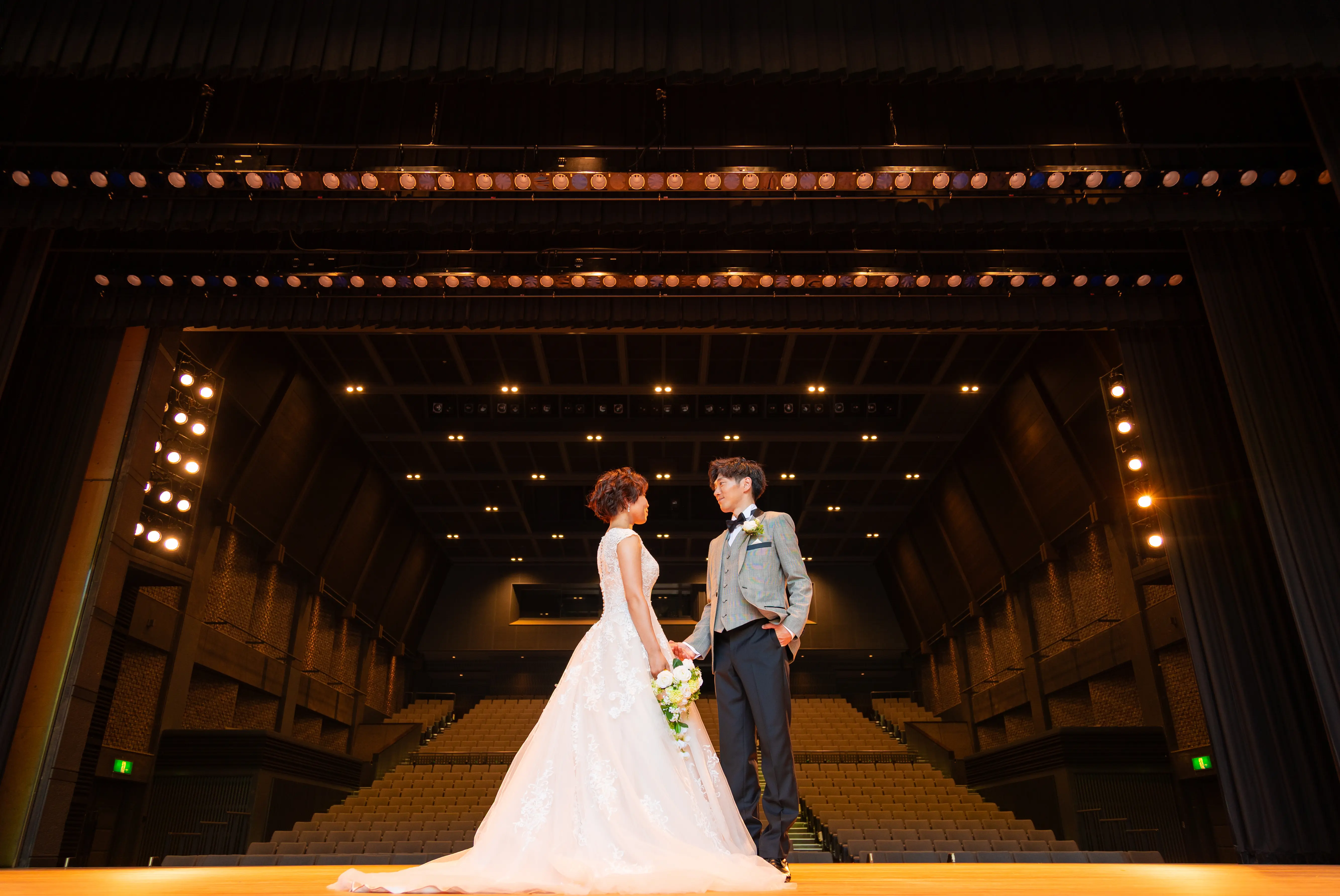 能登演劇堂の舞台上でずらりと並ぶステージ照明のバトンを背に見つめ合う新郎新婦