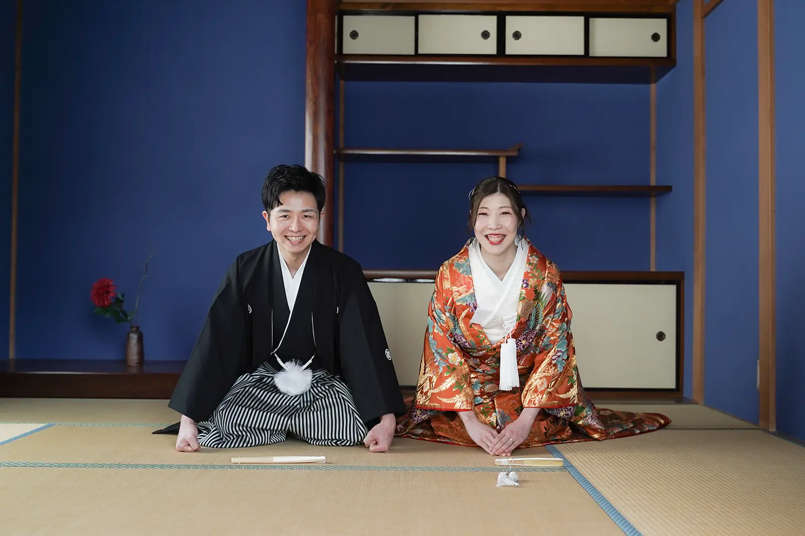 青い和室で正座をして笑顔を見せる和装姿の新郎新婦