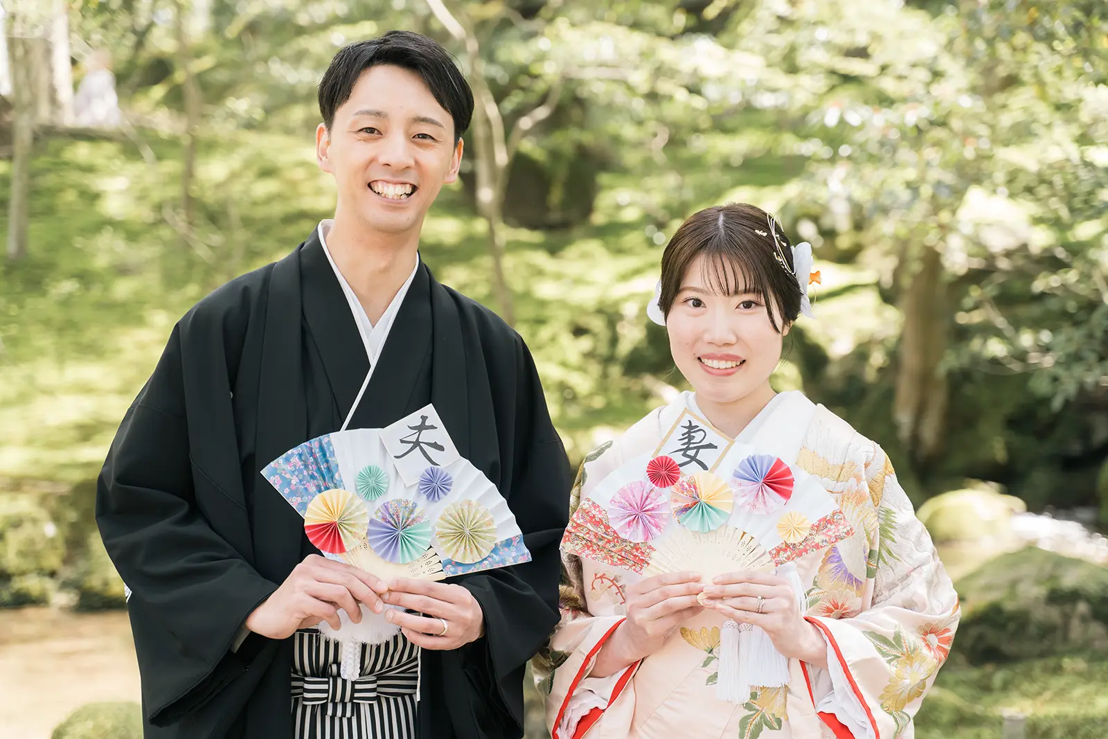 庭園で夫婦扇子を持って笑顔を見せる和装姿の新郎新婦