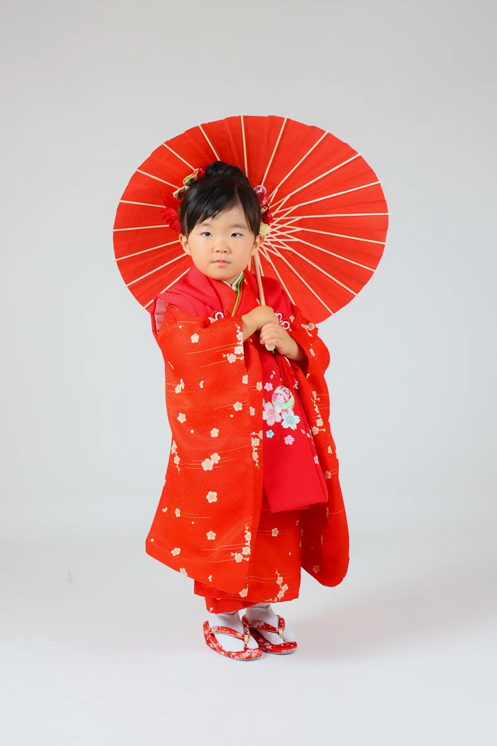 朱色の和傘を持ち白ホリゾントに立つ3歳の女の子、赤い被布姿の七五三スタジオ撮影（金沢店）