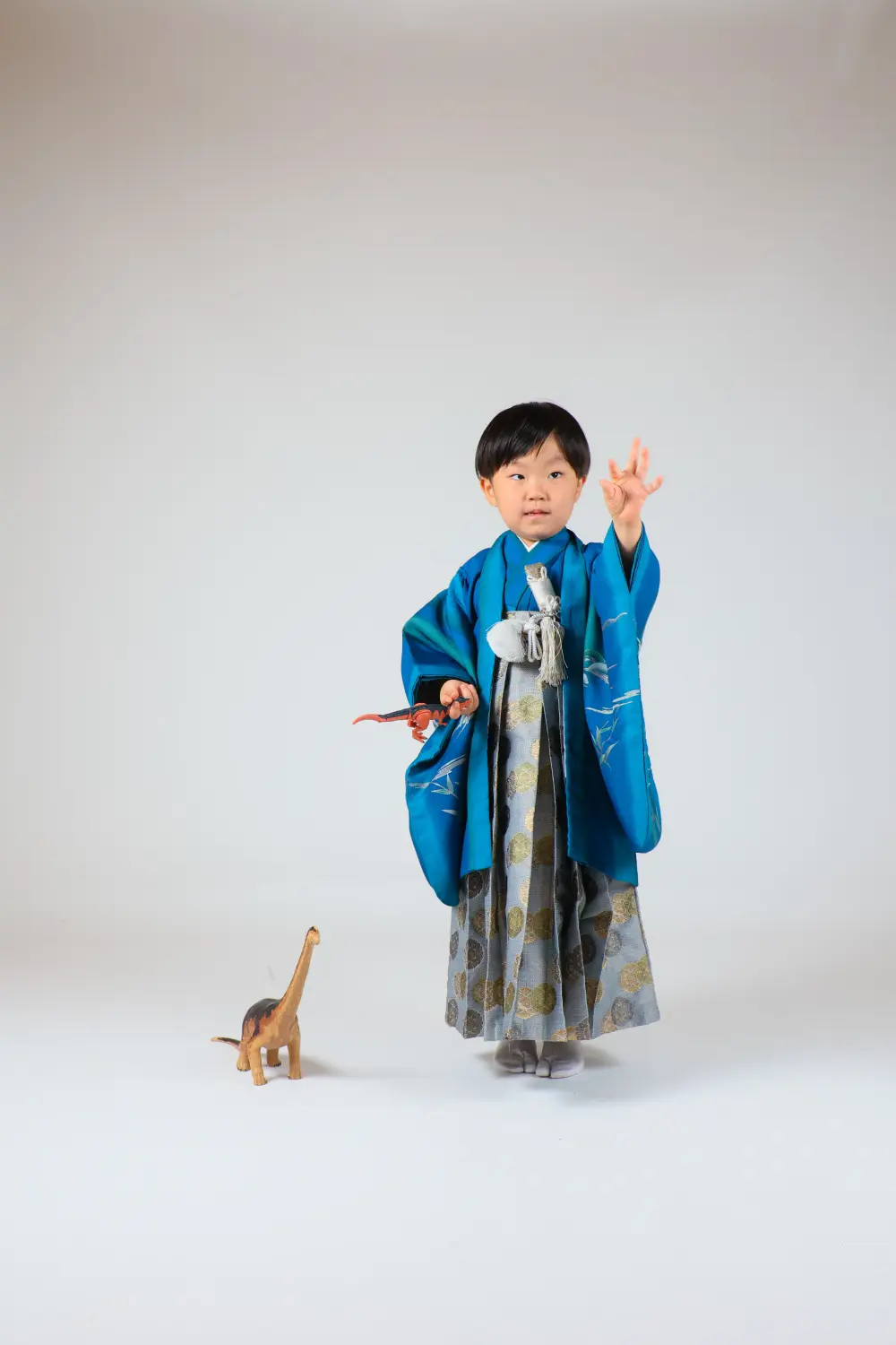 恐竜のおもちゃを手に立つ5歳の男の子、青い羽織と菊丸文の袴の七五三スタジオ撮影（金沢店）