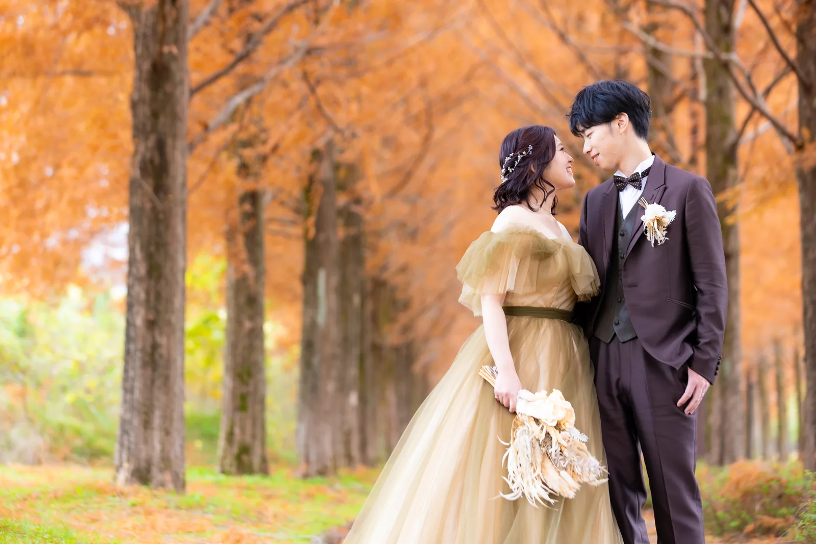 オレンジに色づいたメタセコイア並木で見つめ合う、ゴールドのチュールドレスの新婦とブラウンのタキシードの新郎