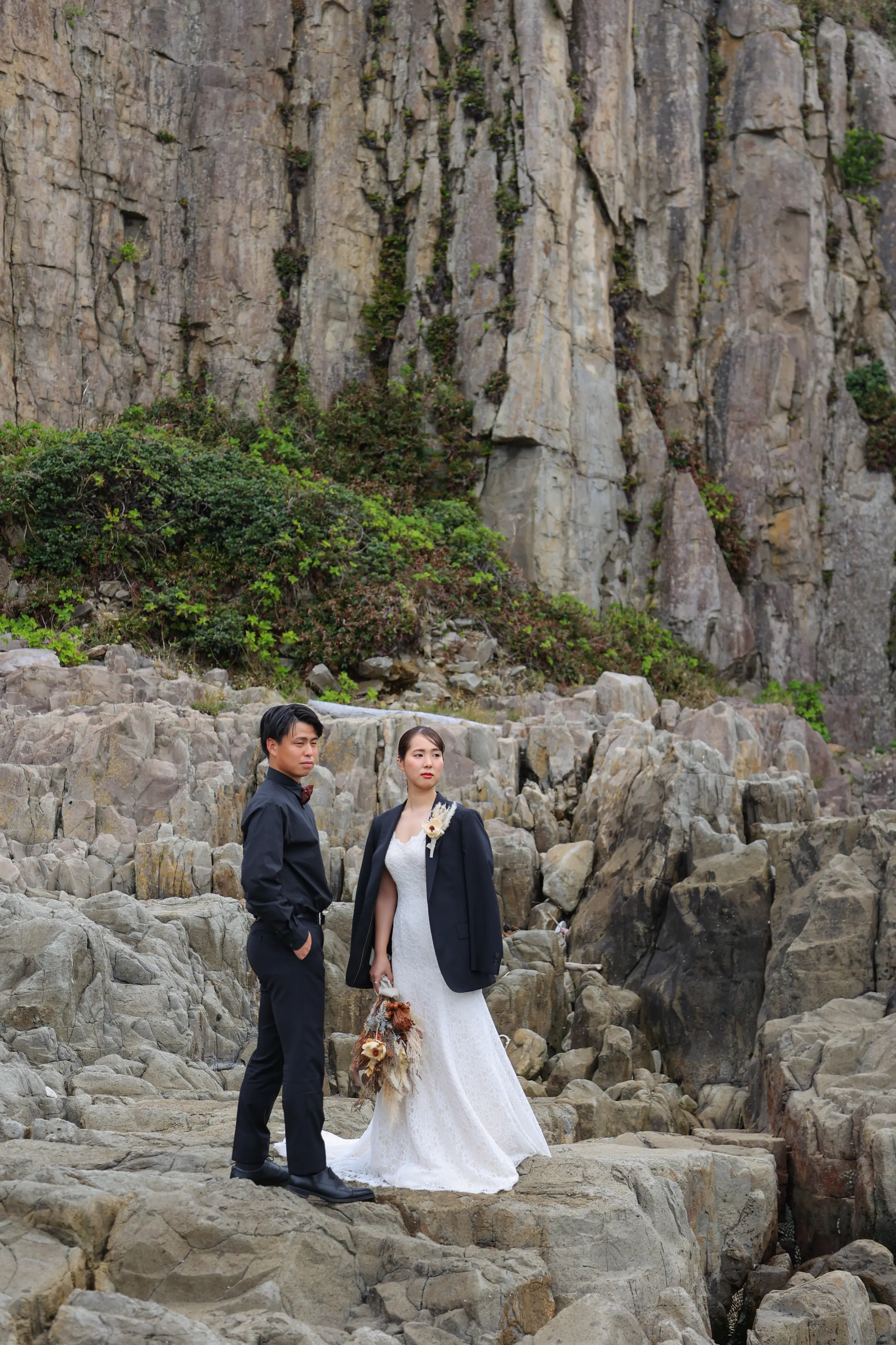 東尋坊の大きな岩壁の前で、ドレスの上に黒いジャケットを羽織った新婦とダークな衣装の新郎が並ぶスタイリッシュな一枚