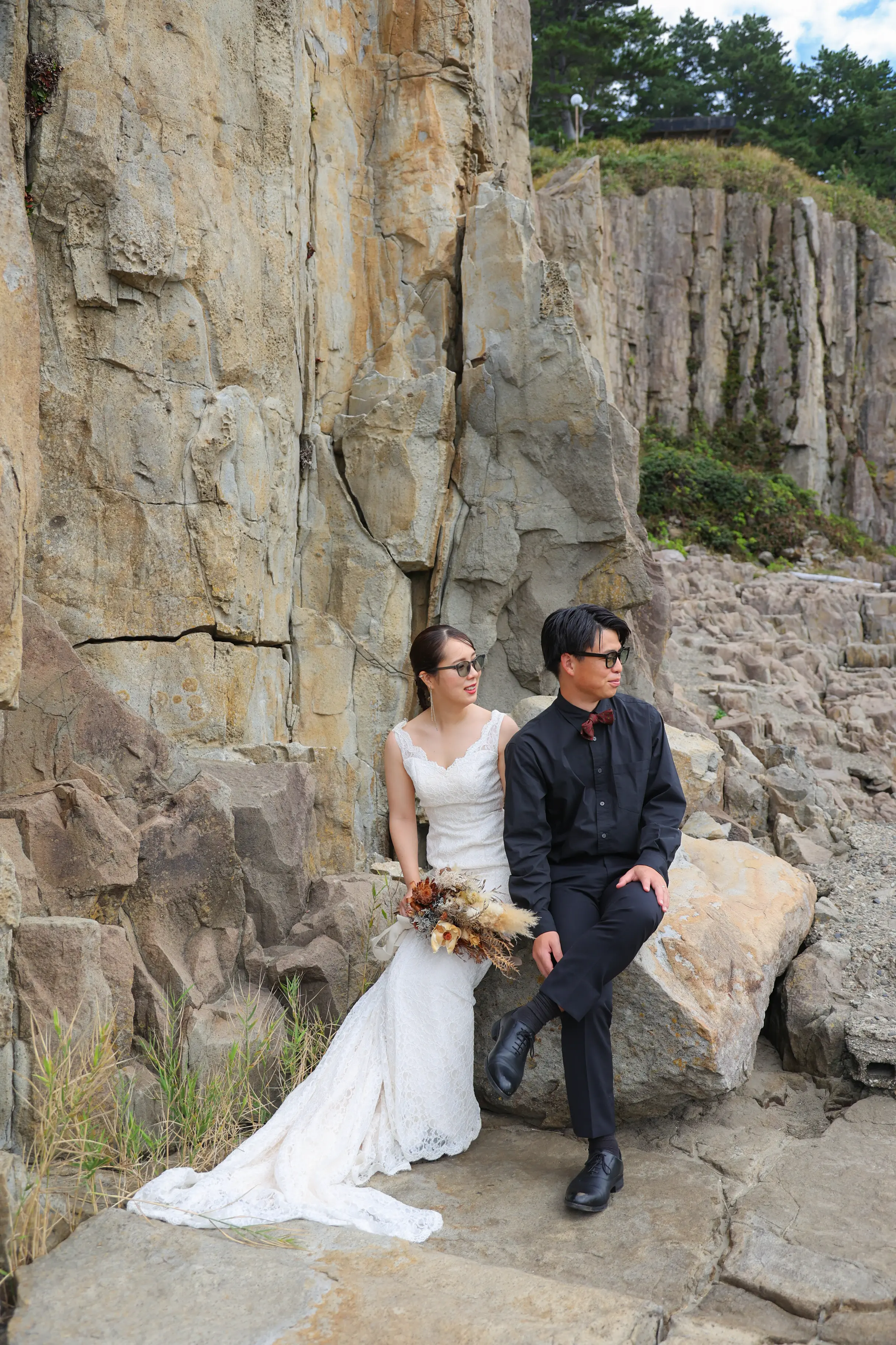 東尋坊の岩に腰かけ、サングラス姿でくつろぐ新郎新婦。青空と松の木が見えるカジュアルな一枚