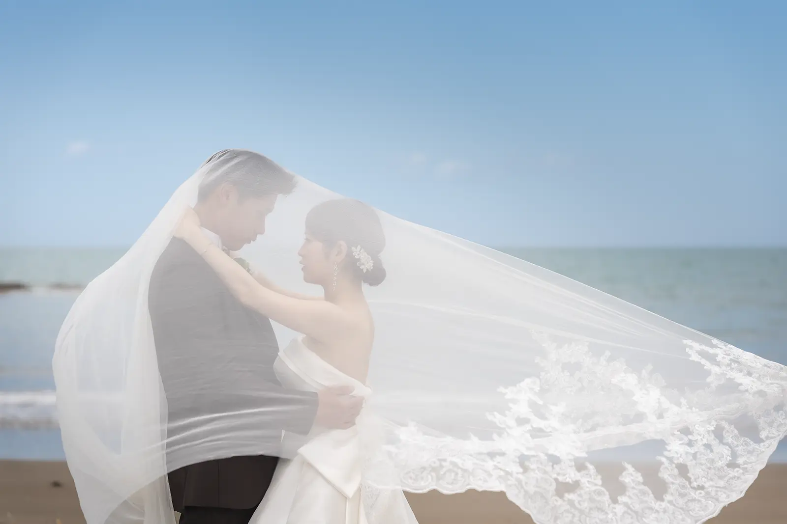 海辺でベールに包まれる洋装の新郎新婦