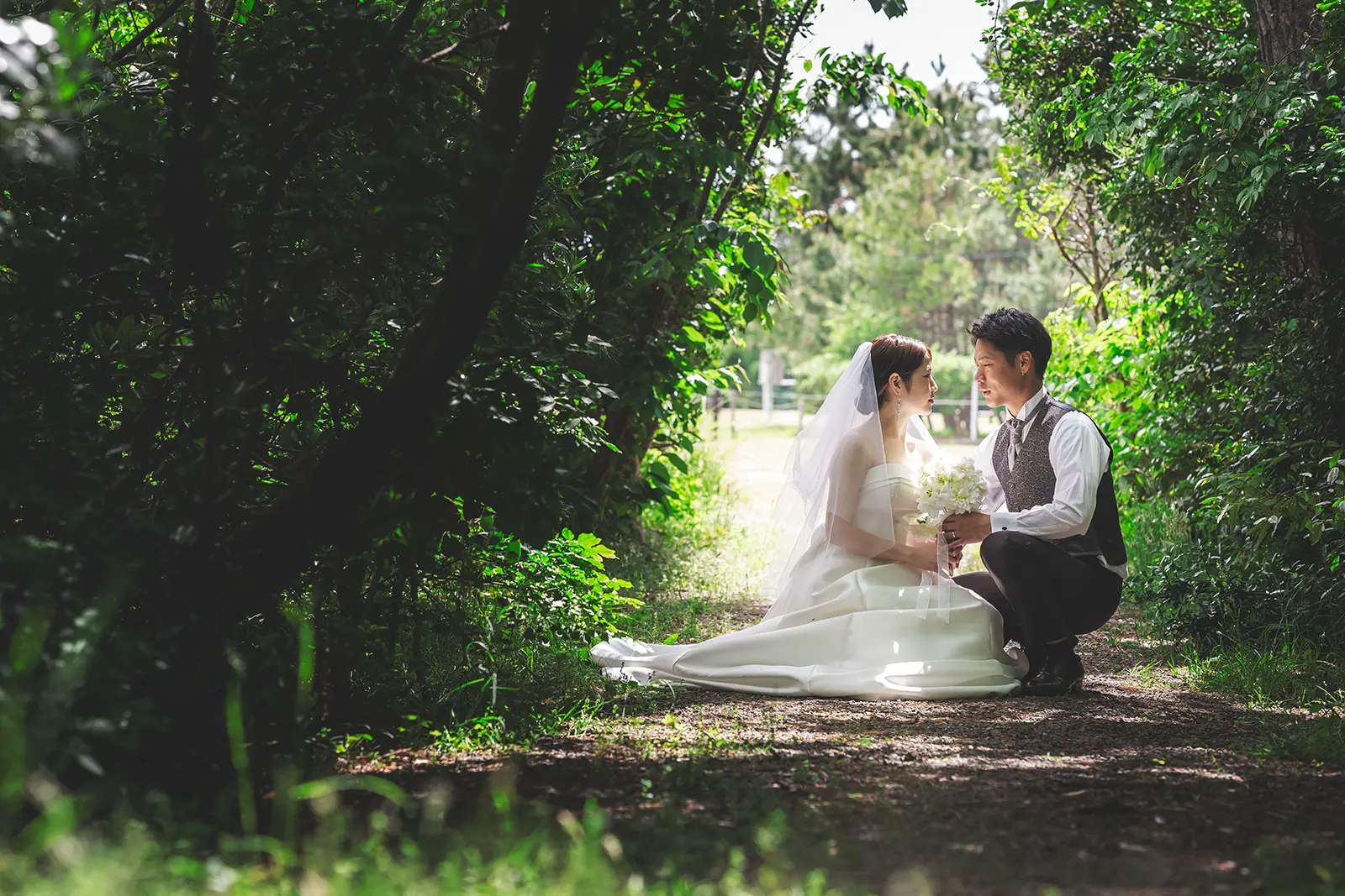 緑に囲まれた小道で向き合う洋装の新郎新婦