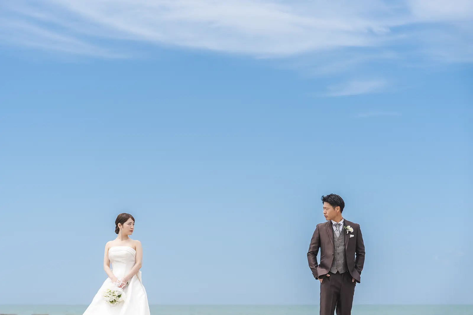 空と海を背景に距離をとって立つ洋装の新郎新婦