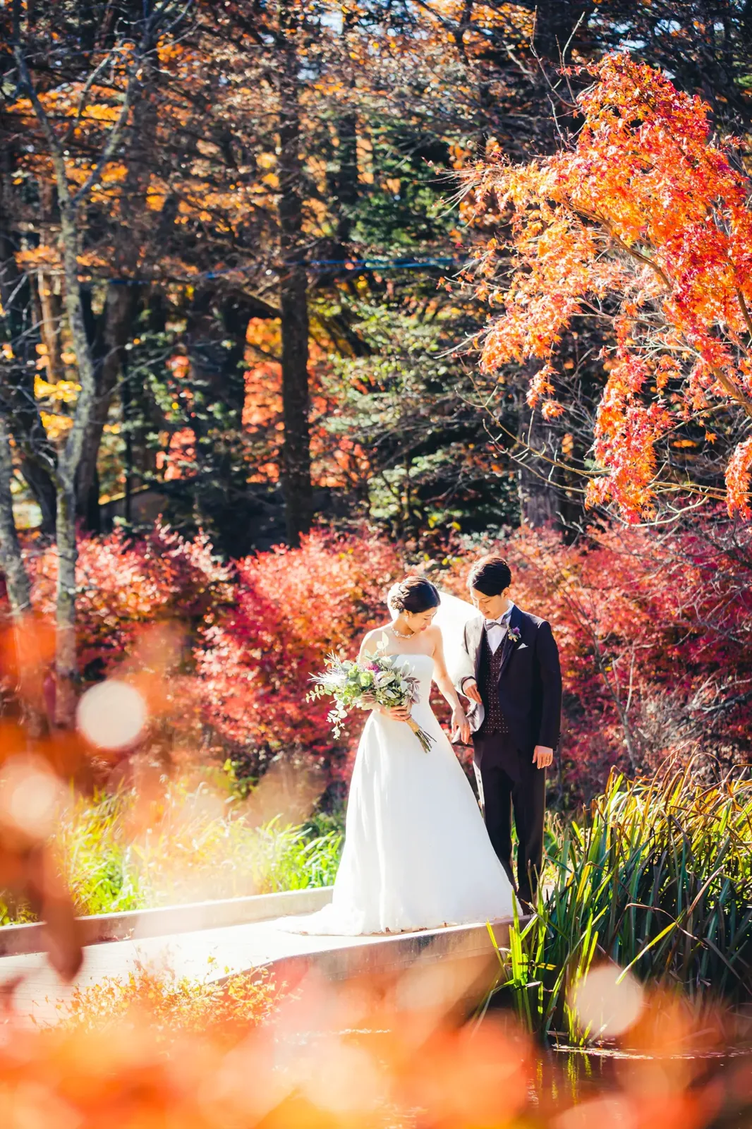 赤い紅葉に囲まれた雲場池の木道で、ブーケを持つウェディングドレスの新婦とタキシードの新郎
