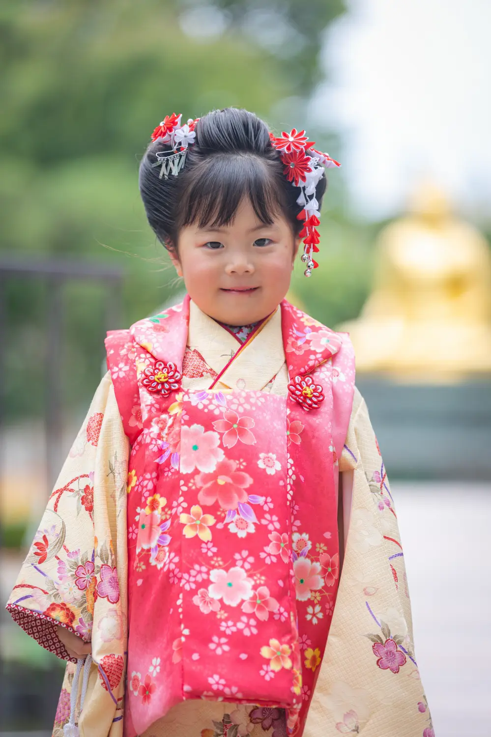 川崎大師の境内で微笑む3歳の女の子、桜柄の赤い被布と日本髪の七五三前撮り（川崎・出張撮影）