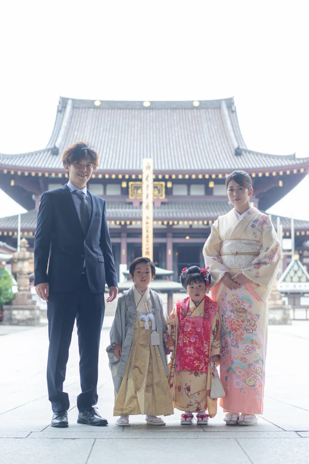 川崎大師の大本堂前に並ぶ家族4人、5歳の羽織袴の兄と3歳の被布の妹、スーツの父と訪問着の母の七五三記念写真