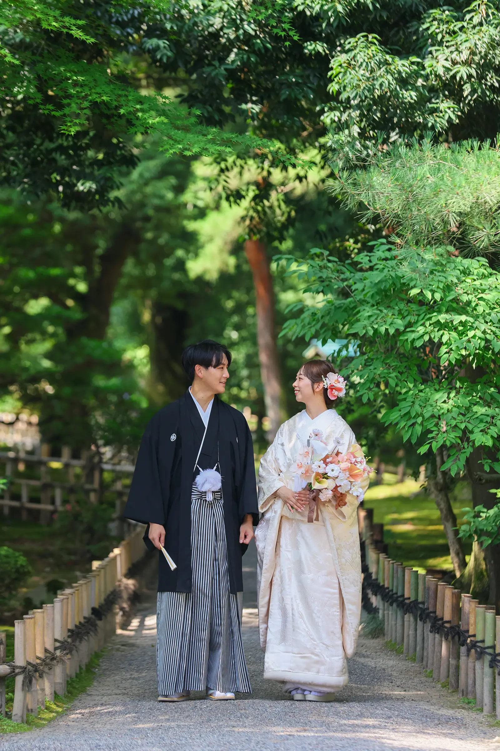 兼六園の新緑に囲まれた小径で白無垢姿の新婦と紋付袴姿の新郎が歩く和装写真