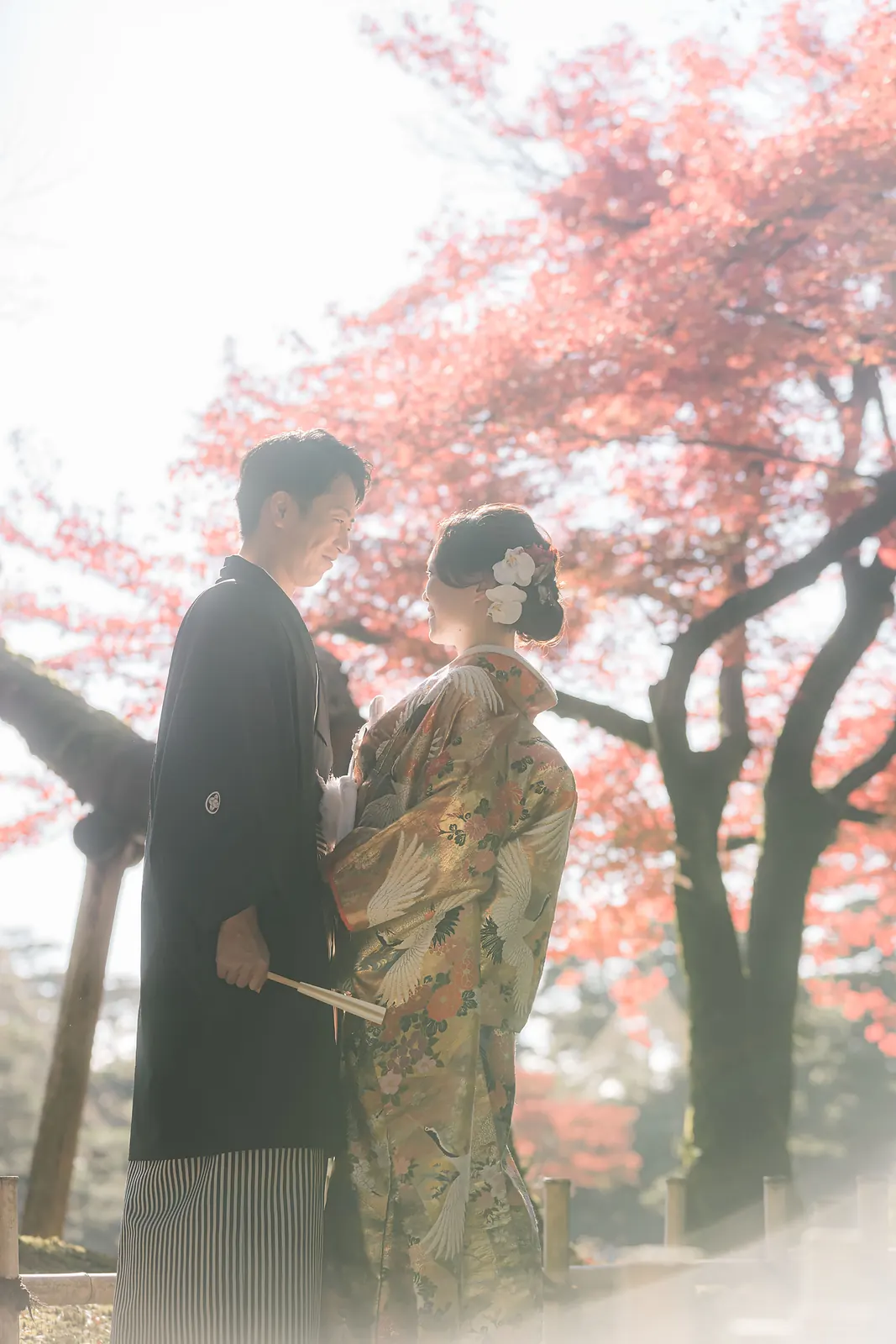 兼六園の紅葉を透かす逆光の中で、向かい合って見つめ合う色打掛の花嫁と紋付袴の新郎