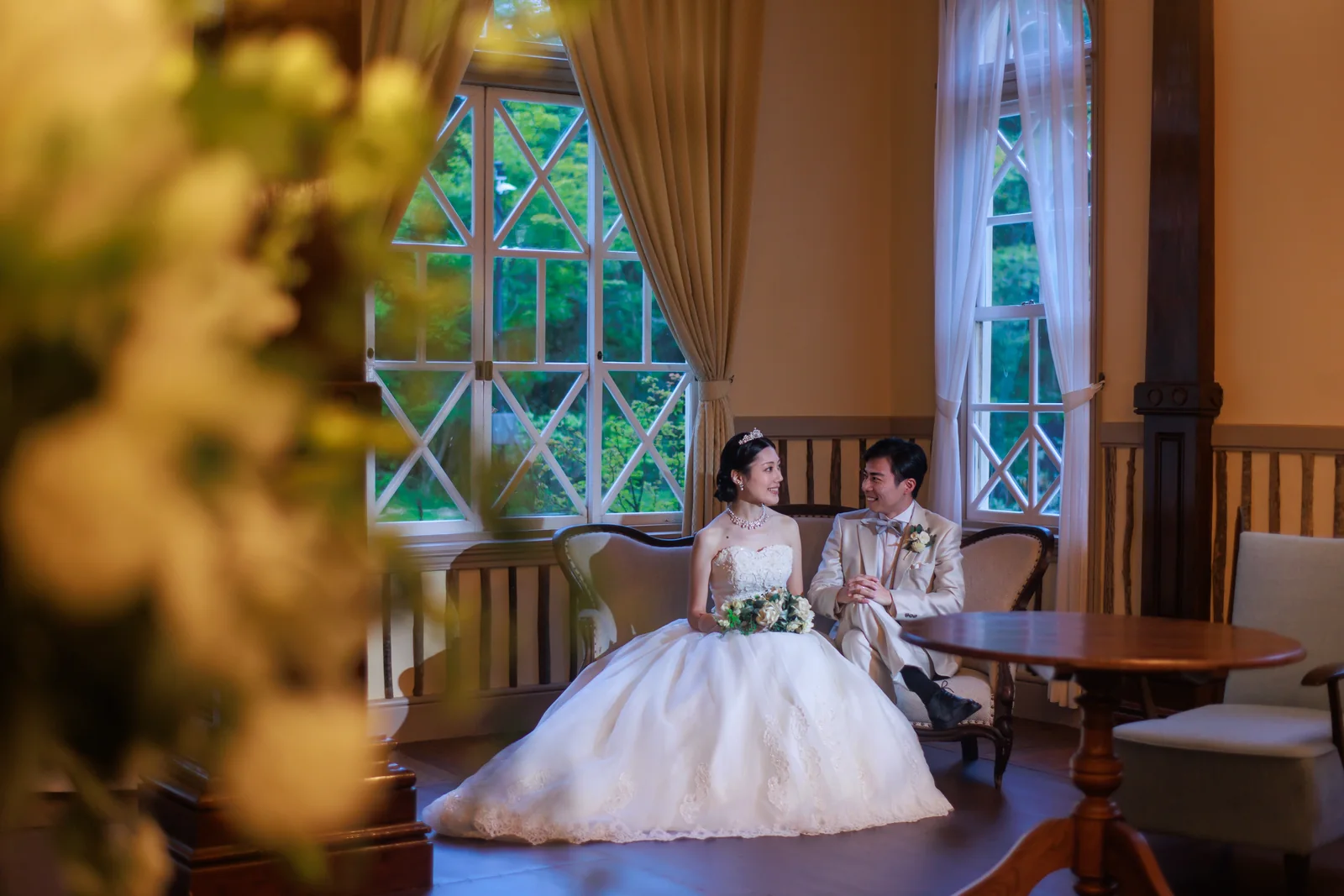 旧三笠ホテルの窓辺のテーブル席に並んで座る新郎新婦を、手前の植栽越しに捉えたカット