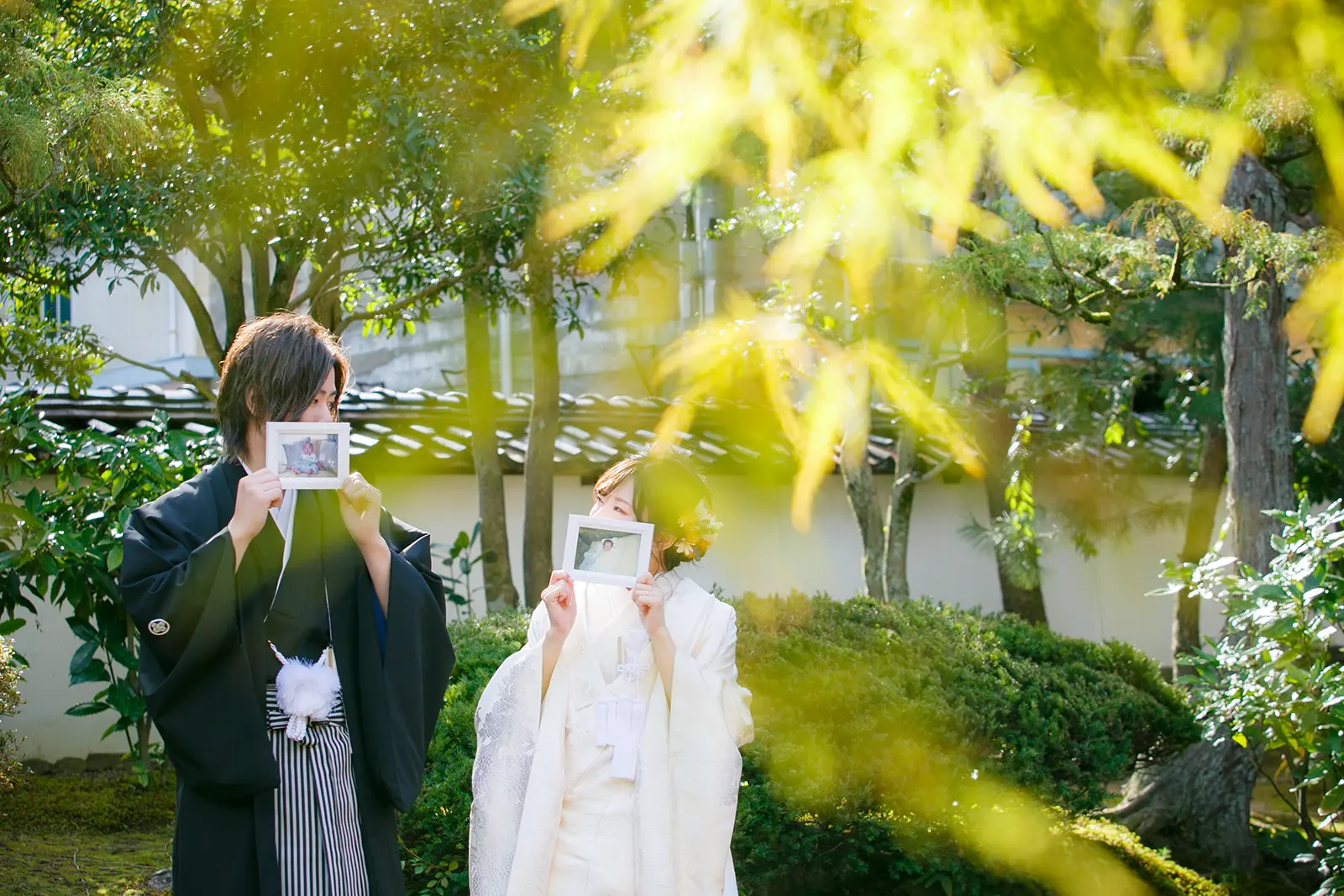 旧高峯家の庭で新郎新婦が幼少期の写真を持って顔を隠す和装前撮り写真