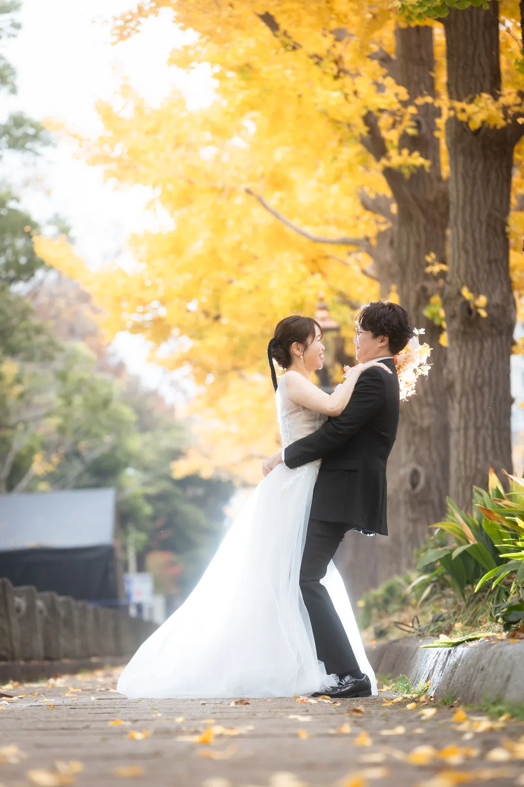 日本大通りのイチョウ並木の下で、ウェディングドレスの新婦を抱き上げるタキシードの新郎