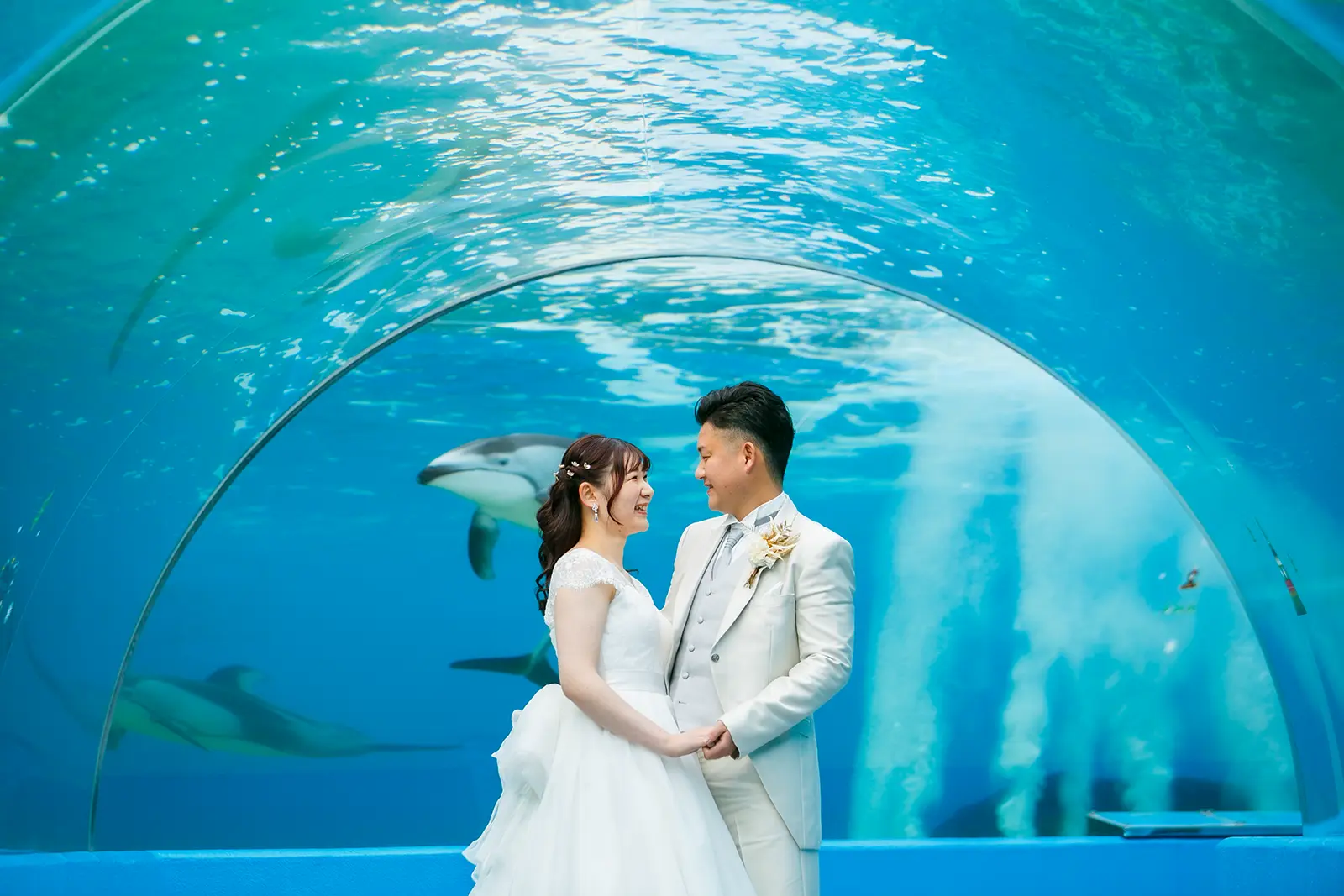 のとじま水族館の青いトンネル水槽の前で向かい合う洋装姿の新郎新婦