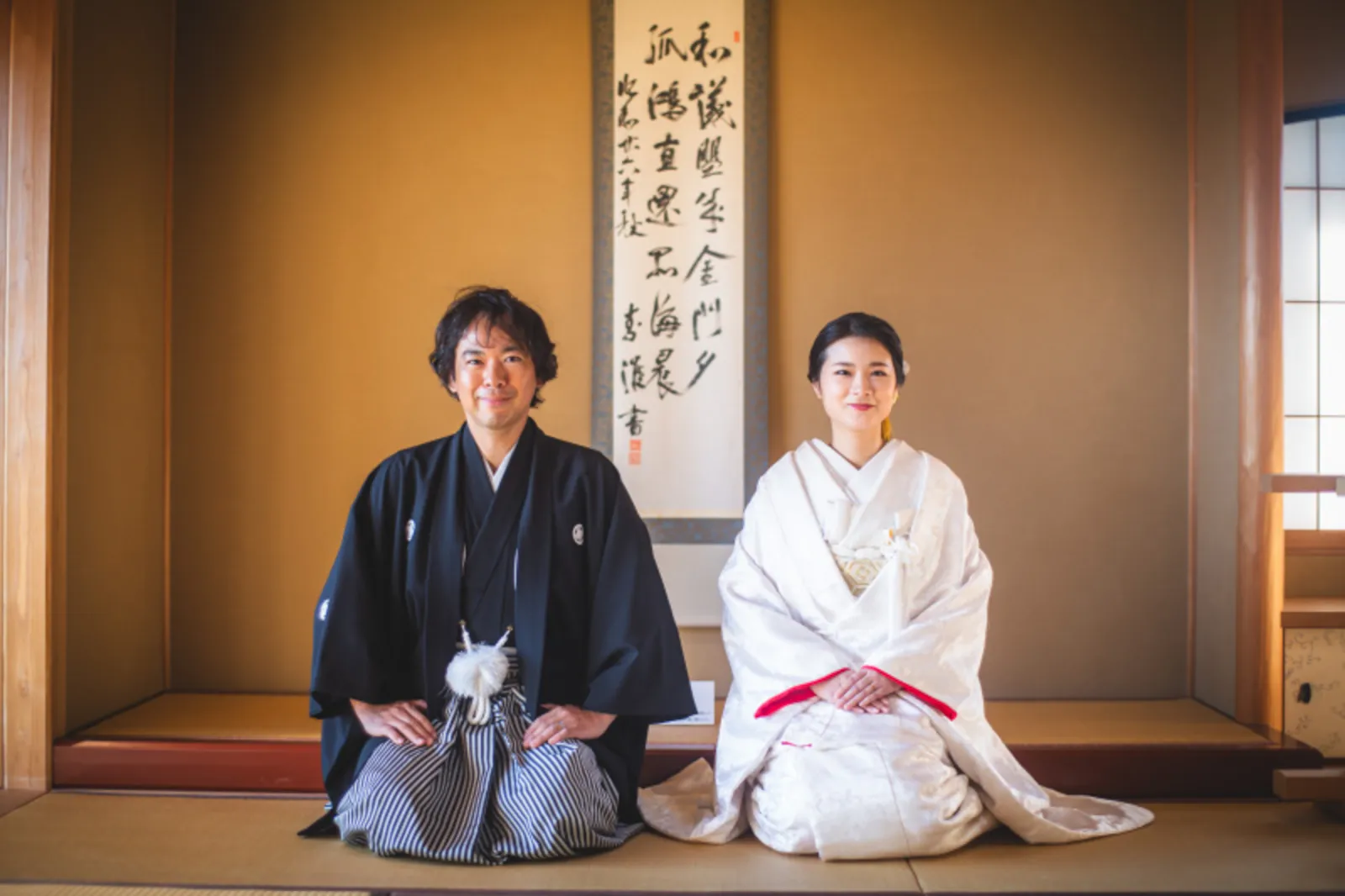 床の間の前で正面を向いて挨拶する和装の新郎新婦の華やかな笑顔