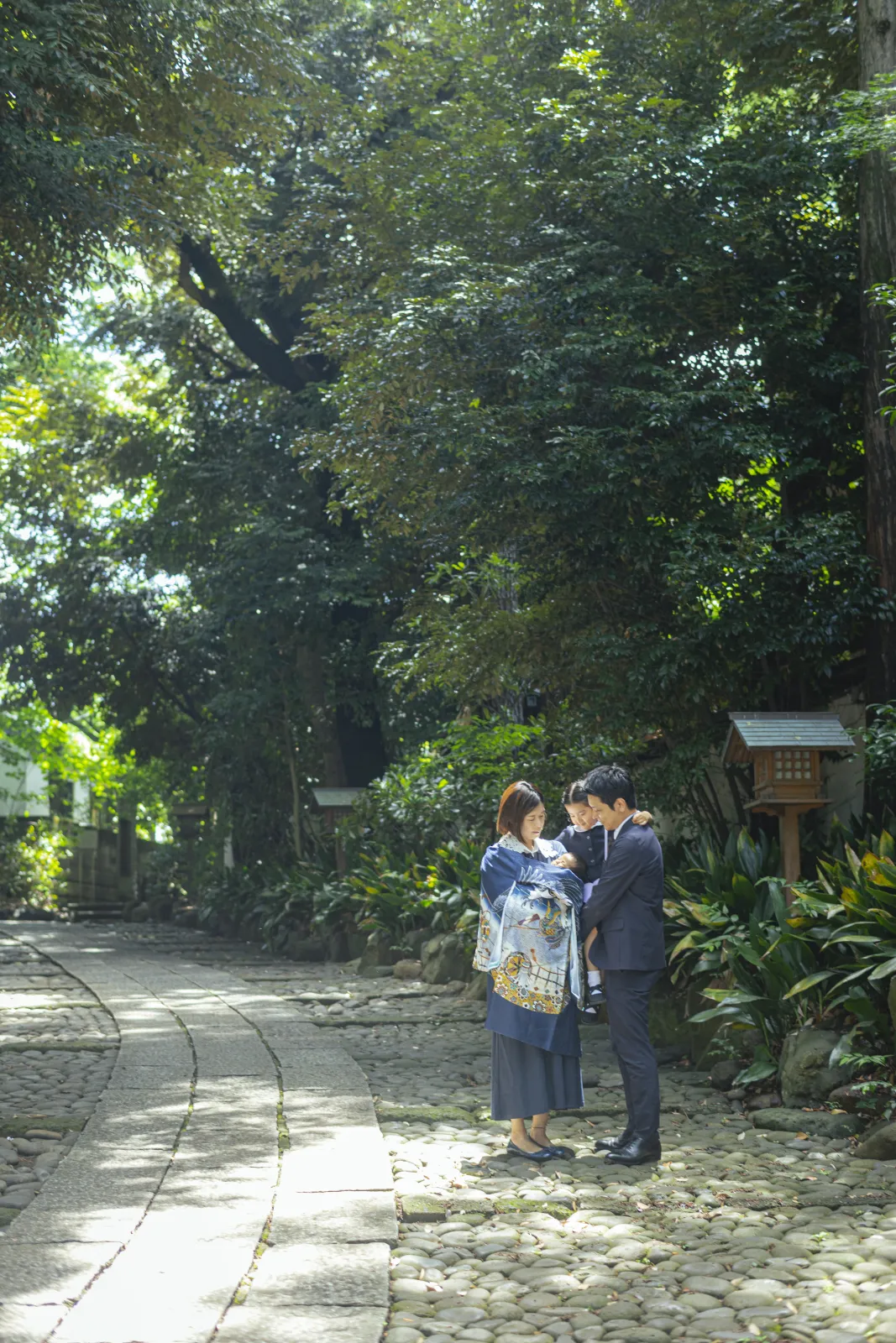 木々に囲まれた神社の参道で、産着を掛けた赤ちゃんを抱く母と、上の子を抱く父が寄り添う引きの家族写真
