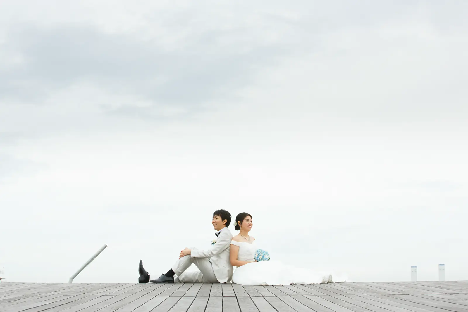 広い空を背景に背中合わせで座る新郎新婦のフォトウェディング写真