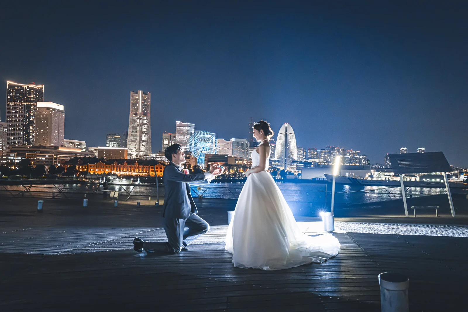夜の大桟橋でみなとみらいの夜景を背景にひざまずいて花嫁の手を取るタキシードの新郎