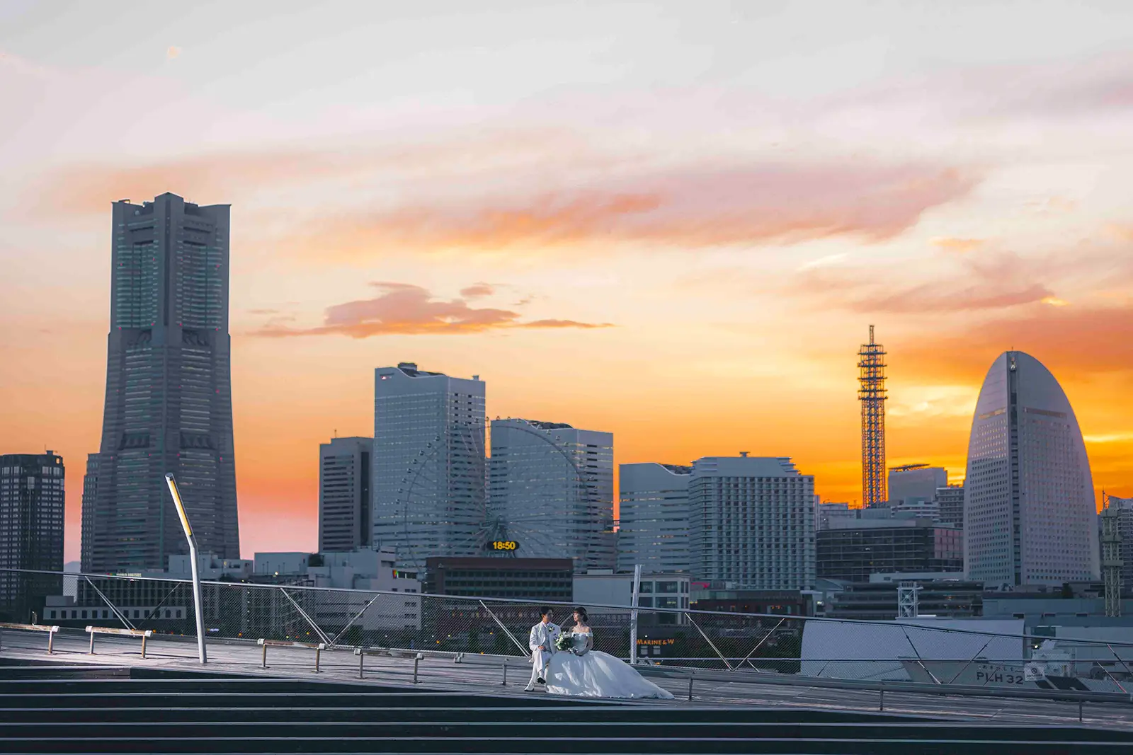 横浜の大さん橋で夕焼けの街並みを背景に座る新郎新婦