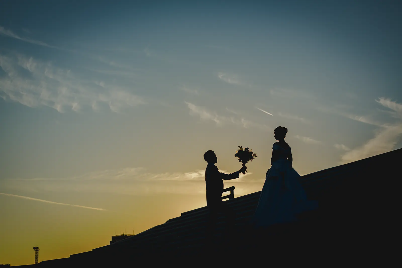 横浜の大さん橋で夕景のシルエットの中ひざまずいて花束を差し出す新郎新婦