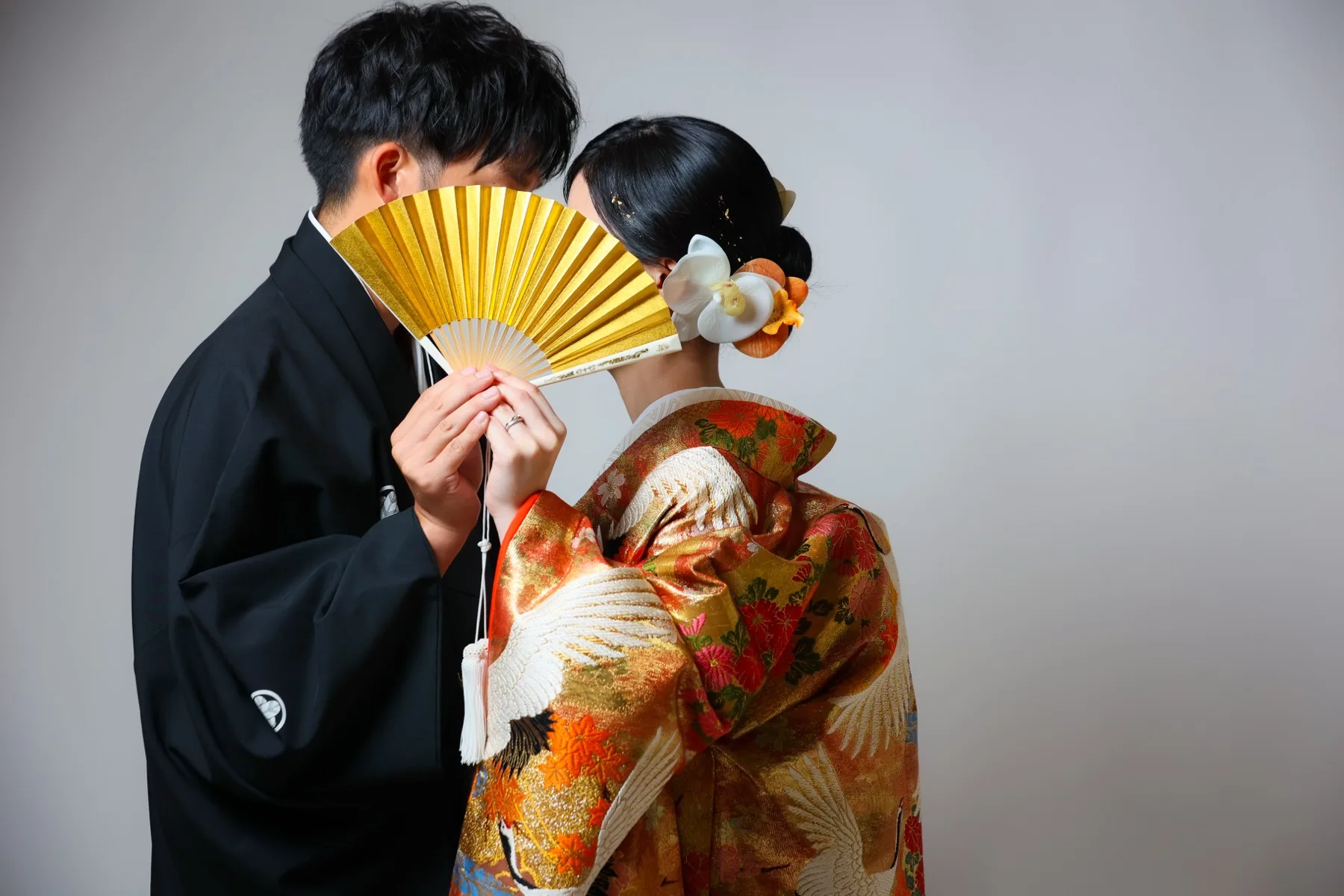金の扇子を手に向かい合う黒紋付の新郎と鶴柄の色打掛の花嫁｜金沢のスタジオ和装撮影