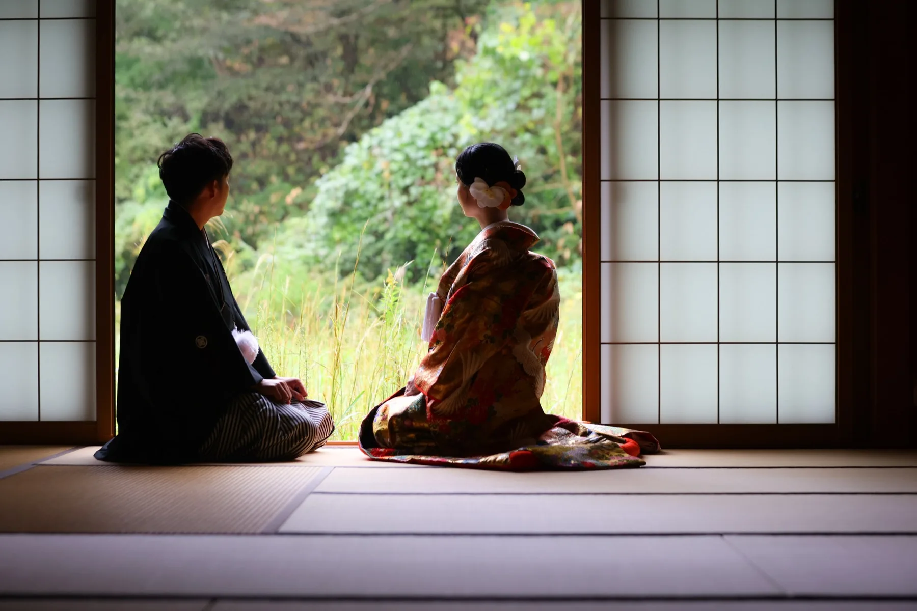障子の和室で庭の緑を眺める色打掛の花嫁と黒紋付の新郎の後ろ姿｜金沢のスタジオ和装撮影