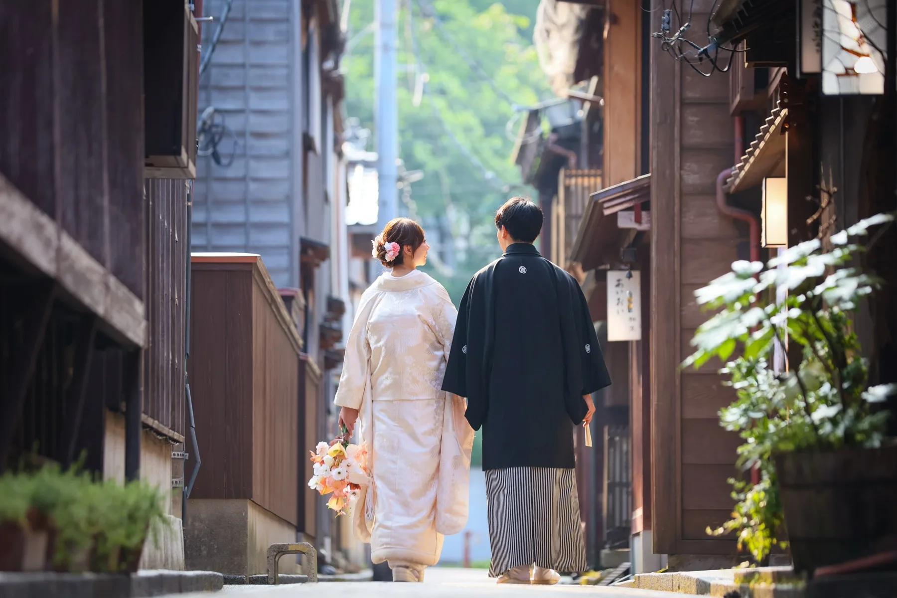 情緒ある街並みの路地を歩く白無垢の花嫁と黒紋付袴の新郎の後ろ姿、オレンジのブーケ｜金沢の和装ロケーションフォトウェディング