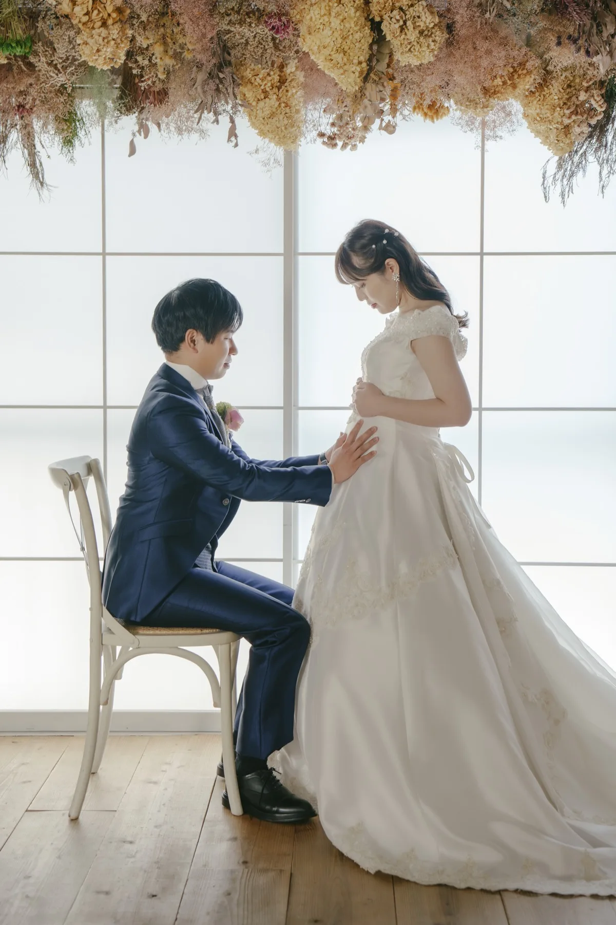 ドライフラワーが飾られた横浜のスタジオで、新郎が花嫁のお腹にそっと手を添えるマタニティフォトウェディング｜フォトジェリッシュ横浜