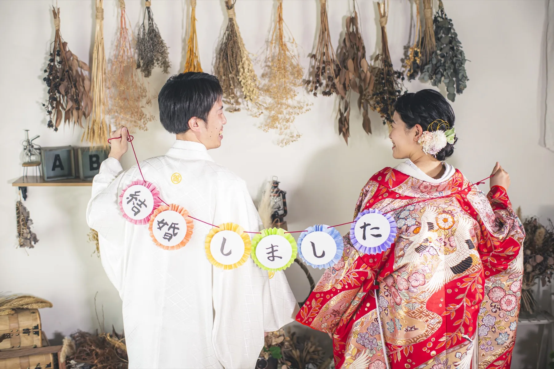 ドライフラワーのスタジオで「結婚しました」のガーランドを持ち見つめ合う、赤い色打掛の花嫁と白い紋付の新郎｜横浜の和装フォトウェディング