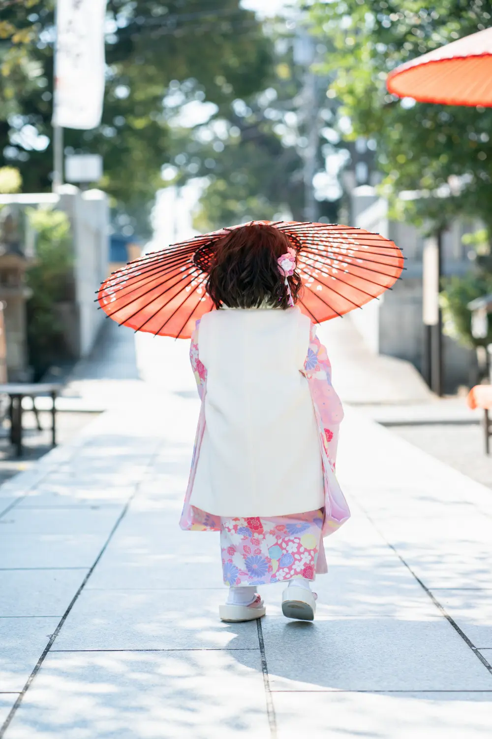 朱色の和傘を差して六所神社の参道を奥へ歩く、被布姿の3歳の女の子の後ろ姿（七五三）