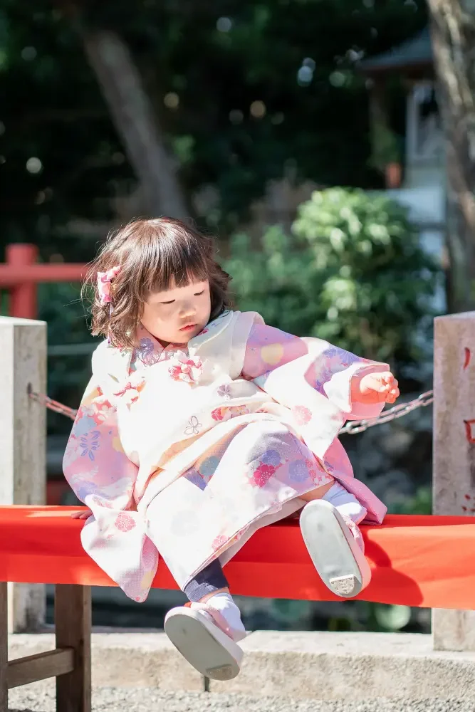赤い布を掛けた縁台に腰かけ、足を投げ出して休む被布姿の3歳の女の子（六所神社の七五三）