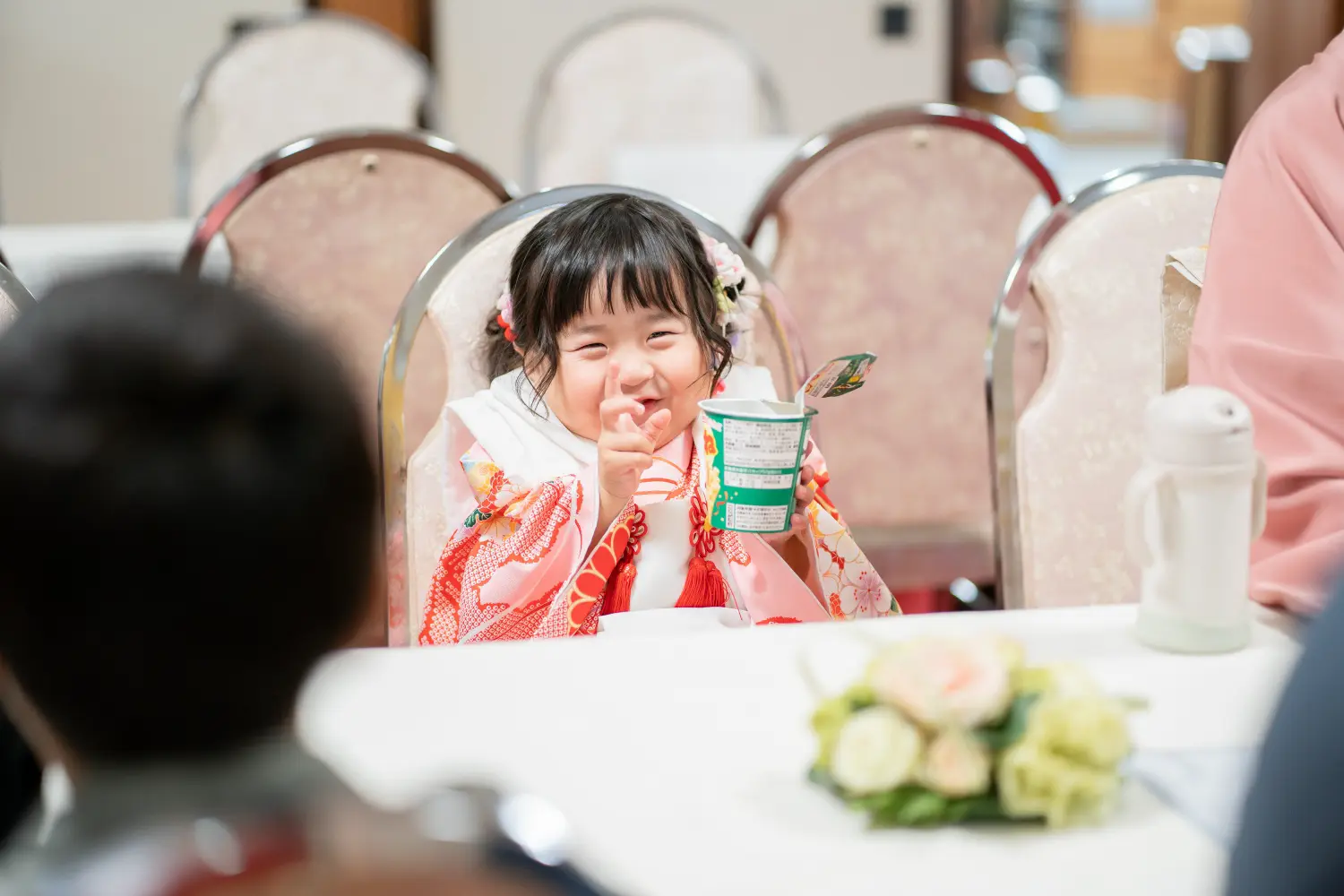 テーブルについてカップを手に笑う被布姿の3歳の女の子、七五三の一日の締めくくり（大磯・六所神社）