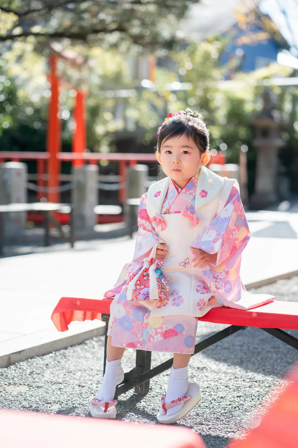 赤い床几に腰かけ足が浮いた被布姿の3歳の女の子、赤鼻緒のぽっくりが見える全身（七五三・六所神社）