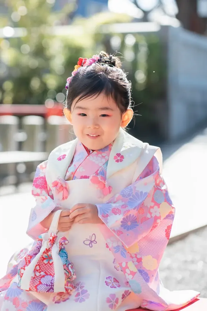 赤い床几に腰かけ巾着を握って照れ笑いする被布姿の3歳の女の子（七五三・大磯）