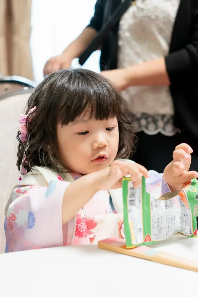 白いテーブルでお菓子の袋を開けようとする、被布姿の3歳の女の子（六所神社の七五三撮影の休憩中）