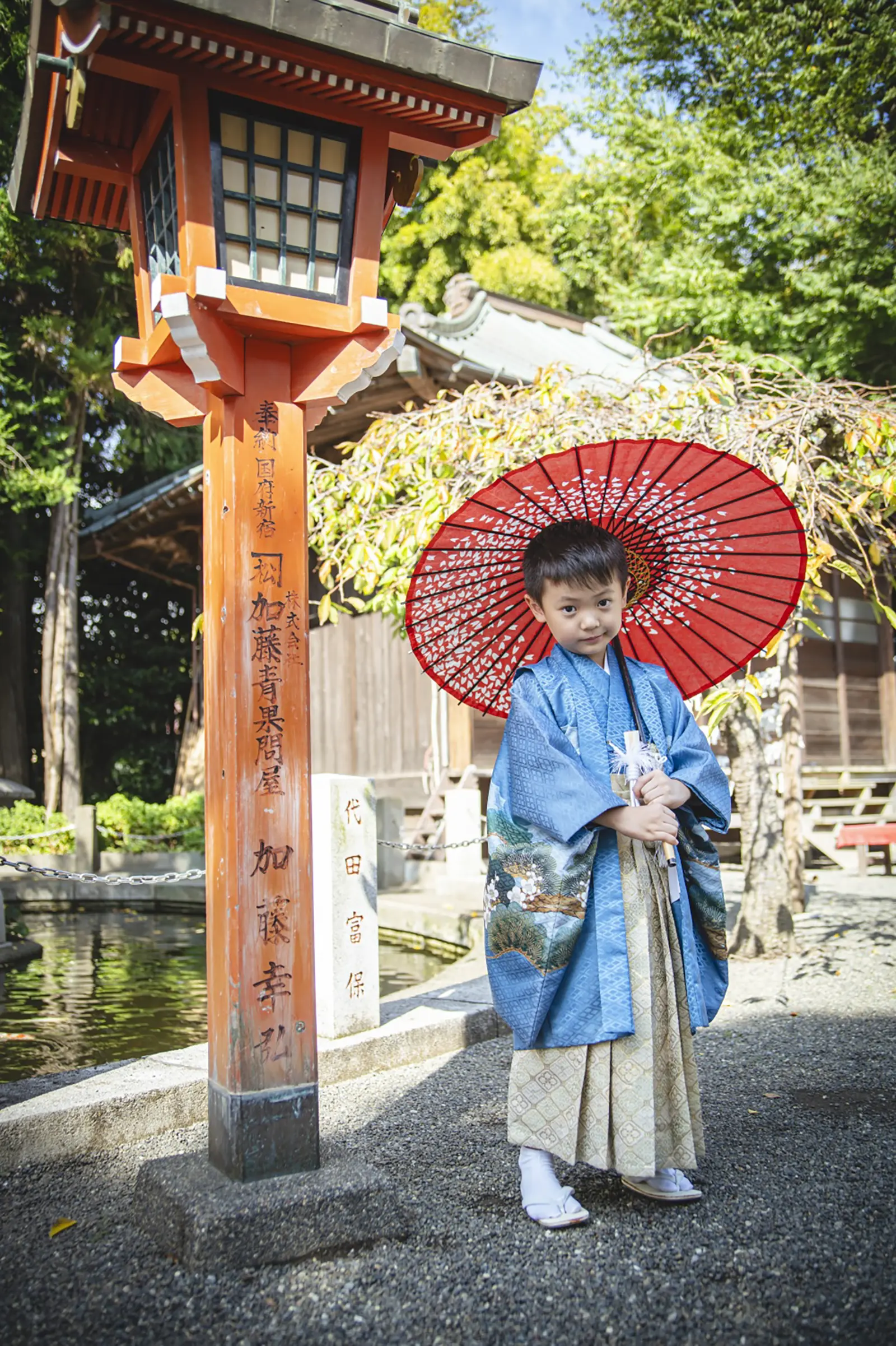 朱の常夜灯と池を背に和傘を手にして立つ5歳の男の子、青い羽織袴の七五三（六所神社・大磯）