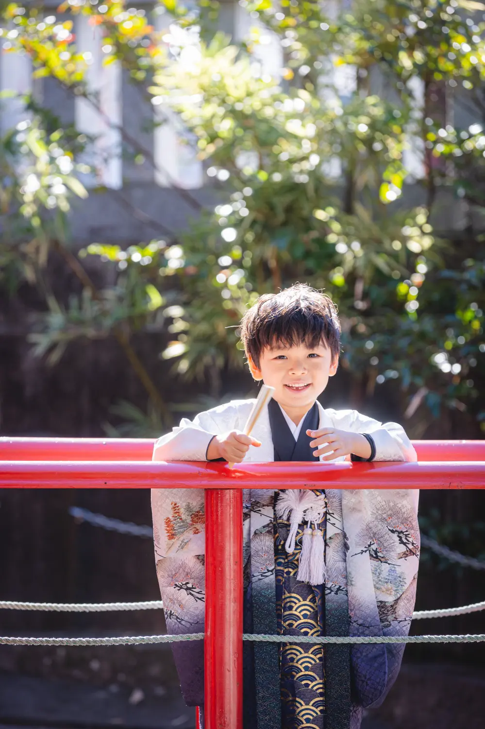 朱塗りの手すり越しに白木の扇を握って笑う5歳の男の子、逆光の緑を背にした七五三（六所神社・大磯）
