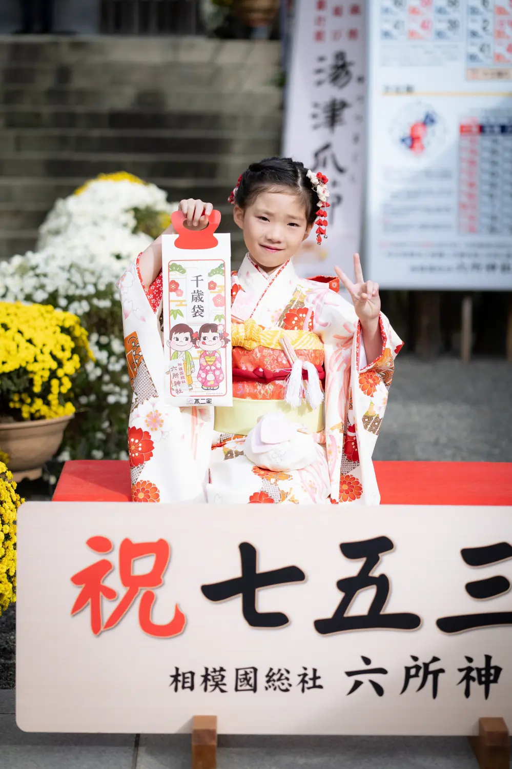 「祝七五三 相模國総社六所神社」の木札の前で千歳袋を掲げる7歳の女の子、四つ身の七五三