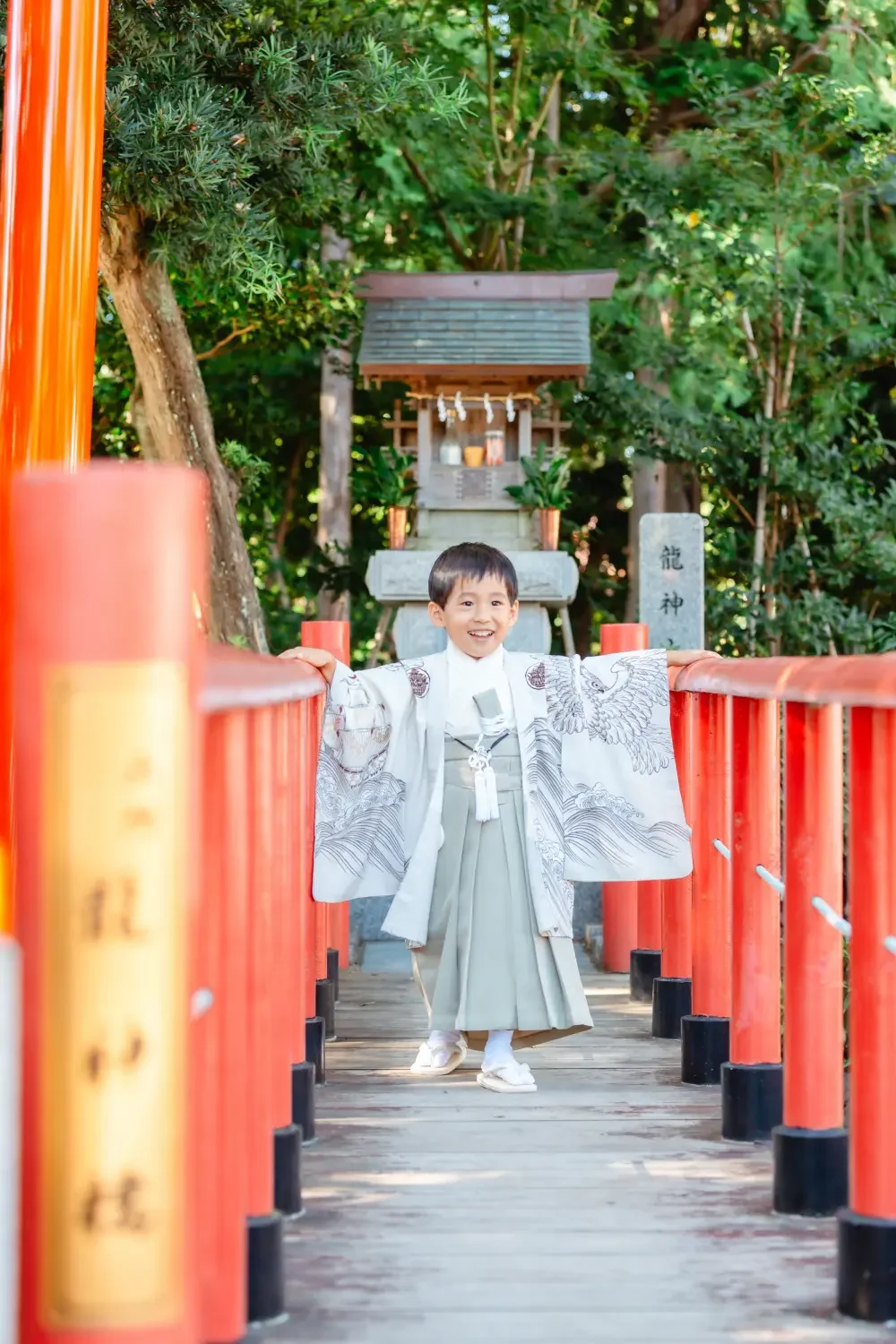 朱塗りの太鼓橋の中央で両袖を翼のように広げて笑う、生成りの羽織袴の男の子の全身、七五三出張撮影（六所神社・大磯）