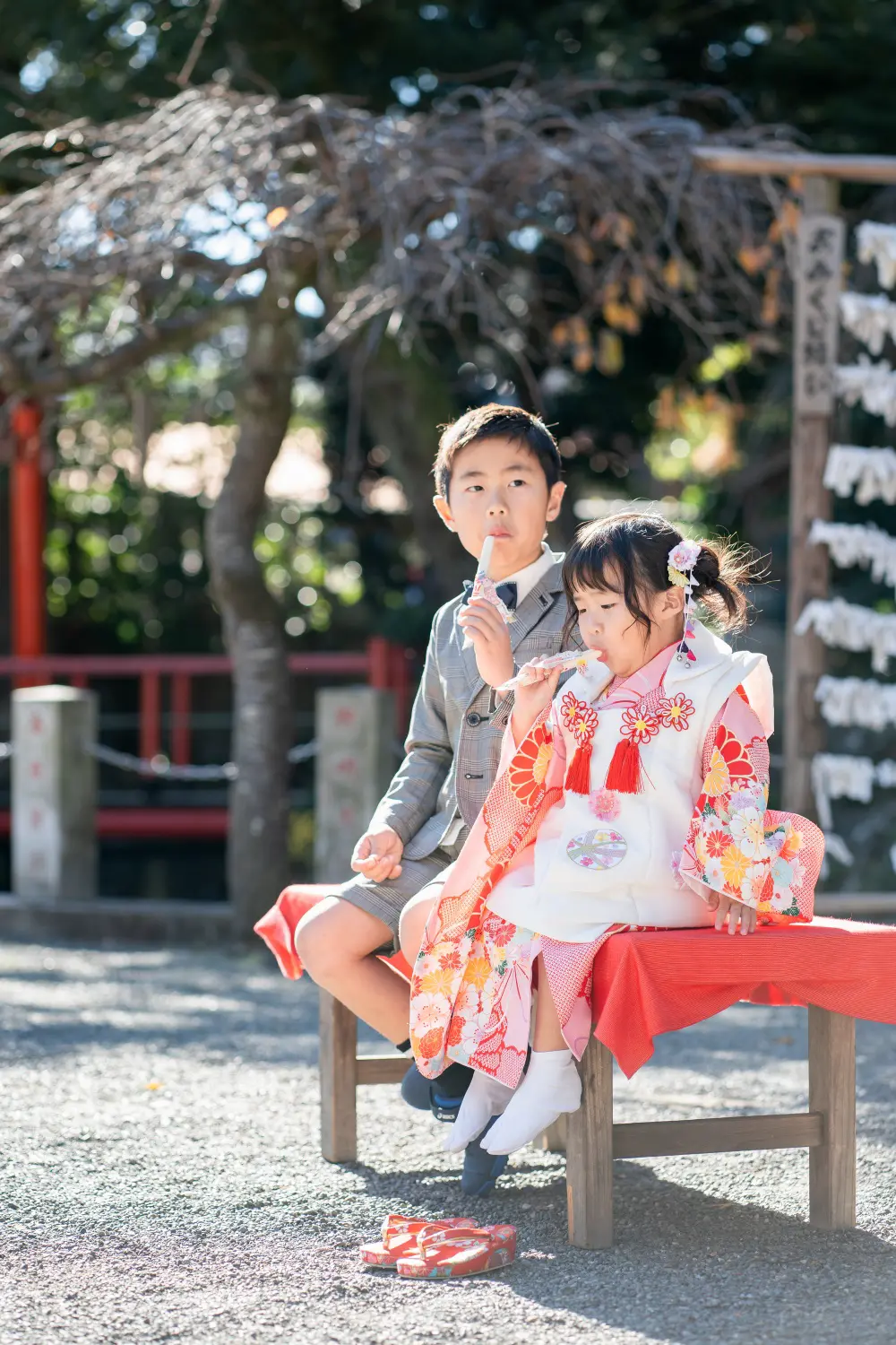 赤い布の縁台に腰かけてお菓子を口にする被布姿の3歳の女の子と兄、六所神社の七五三（大磯）