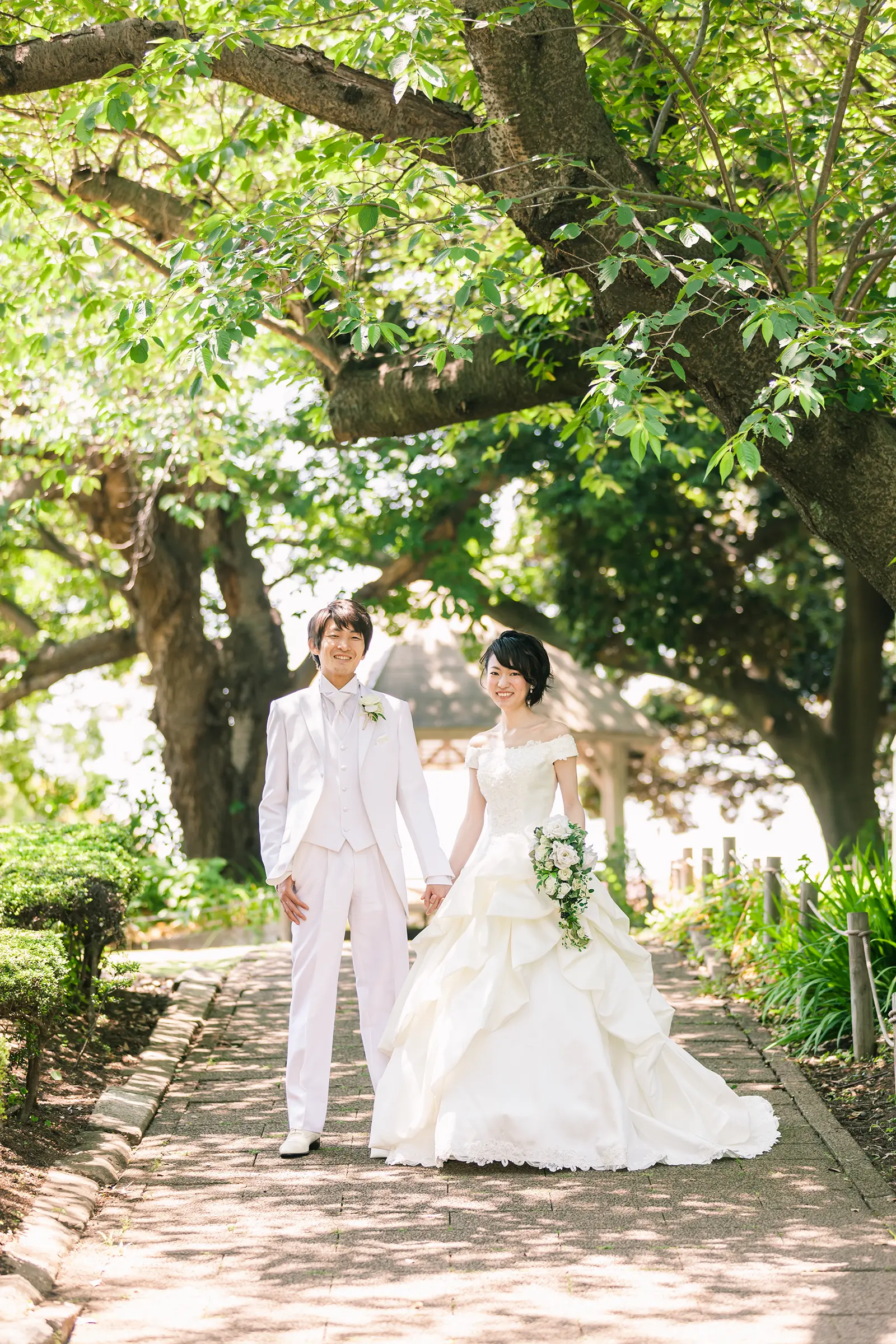 木漏れ日の中で手をつなぐ新郎新婦の全身フォトウェディング写真