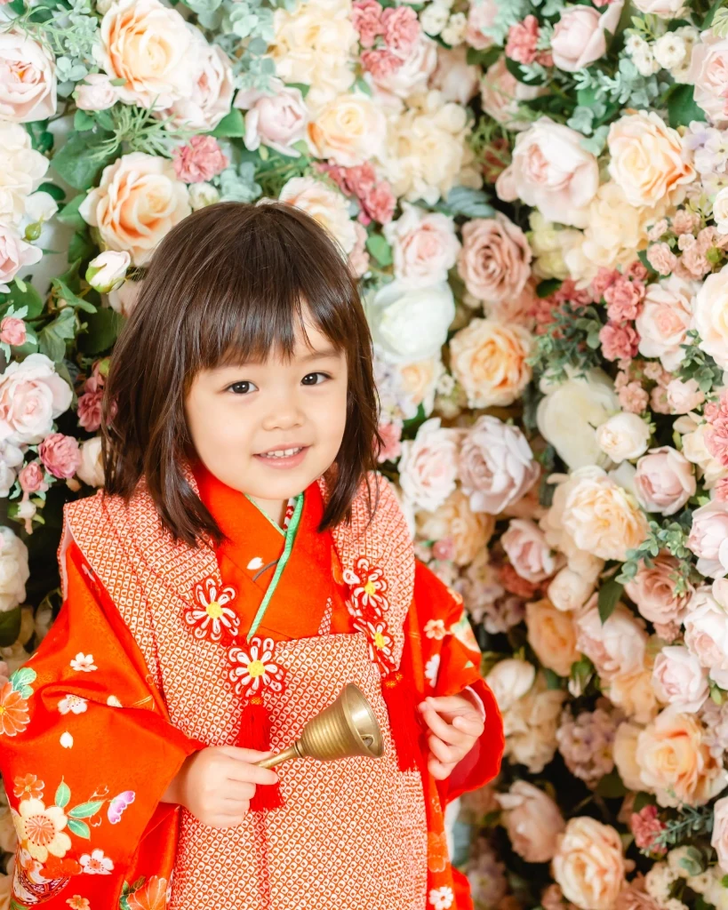 前髪ありのまっすぐなボブを結ばずにおろした3歳の女の子。赤い被布姿で花の壁の前に立つ