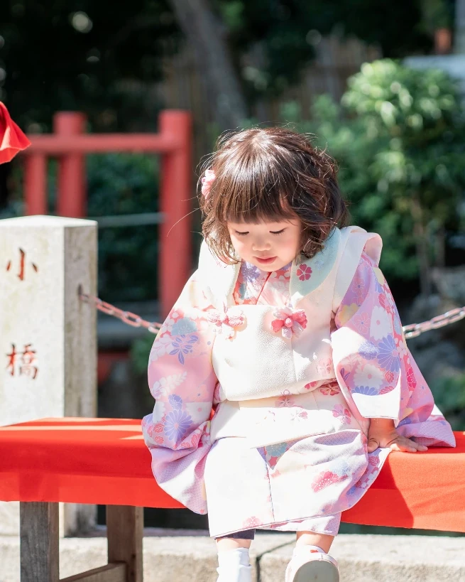 くせ毛のボブをおろしたままの3歳の女の子が、朱色の縁台に座って足もとを見ている